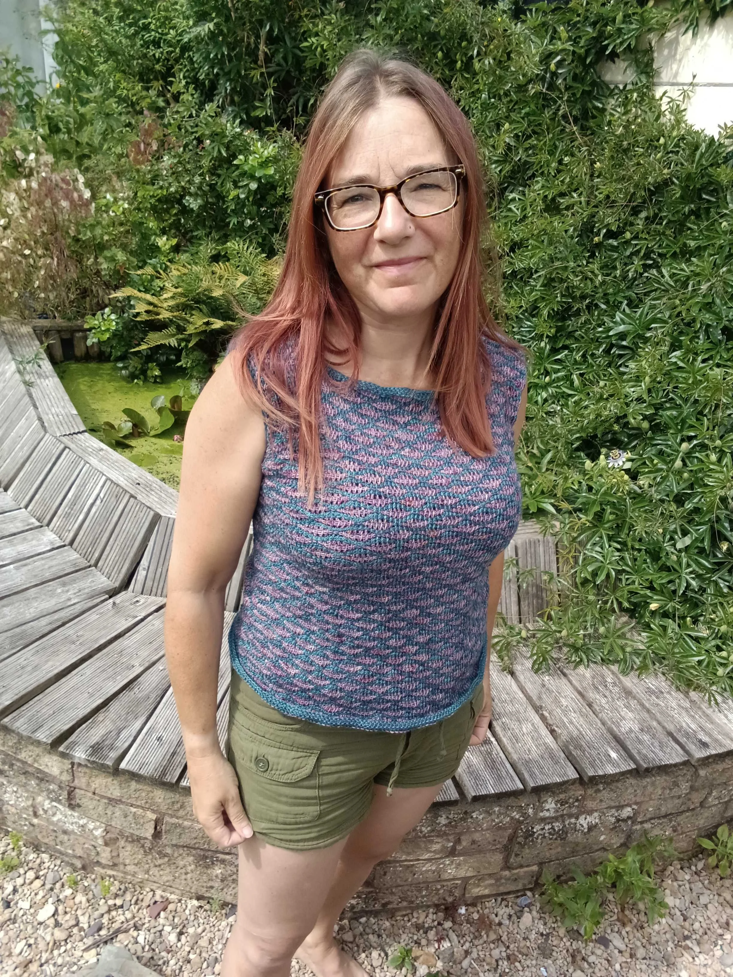 A woman with red hair, glasses, and a nose piercing standing outdoors in front of greenery, wearing a multicoloured sleeveless top and green shorts.