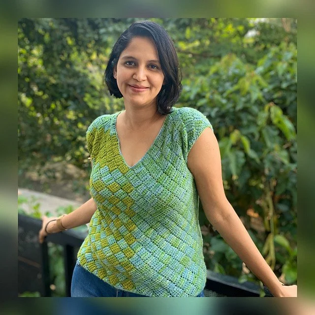 A woman with short black hair smiling outdoors, wearing a green and blue knit top with a geometric pattern, against a background of greenery.