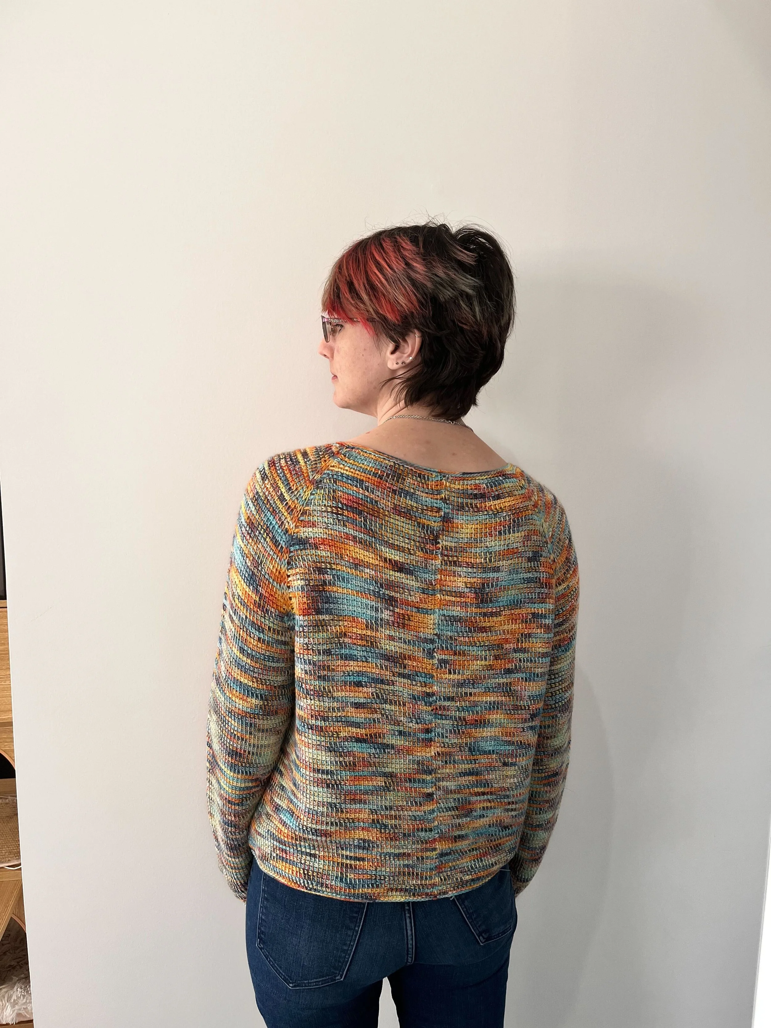 Person with short hair, red highlights, colourful sweater, and blue jeans standing against a plain white wall.