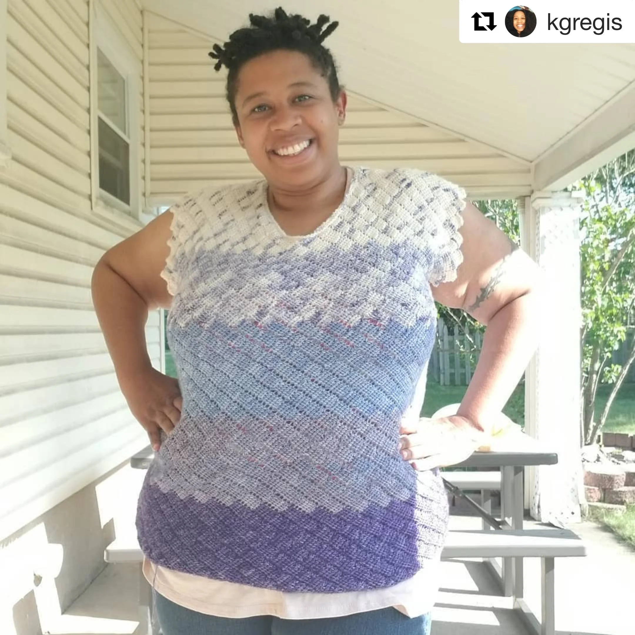 A woman with short curly hair smiling, standing outside on a porch with siding and a window, wearing a textured white top and blue jeans.