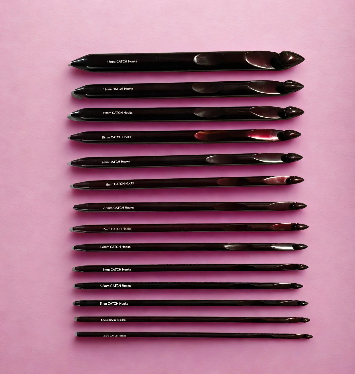 A collection of chocolate coloured Tunisian crochet hooks with different tip sizes, arranged from 15mm to 4.5mm, on a pink background.