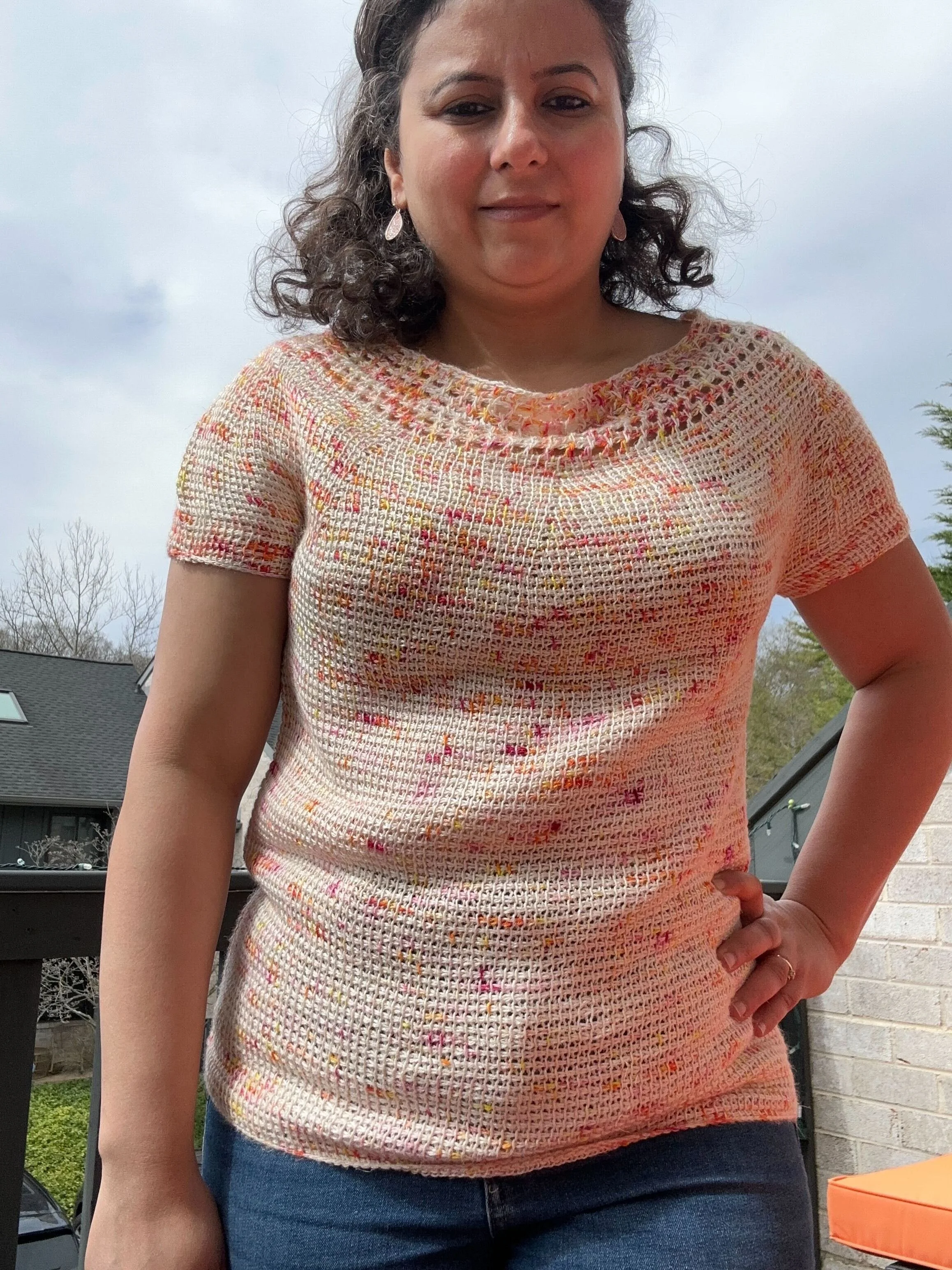 A woman with curly hair wearing a multicolored Tunisian crocheted top and blue jeans standing outdoors near a porch, with a gray house and trees in the background.