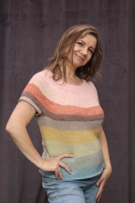 A woman with wavy, shoulder-length hair standing with one hand on her hip and the other at her side, wearing a colorful, striped short-sleeve sweater and jeans, smiling softly at the camera against a dark curtain background.