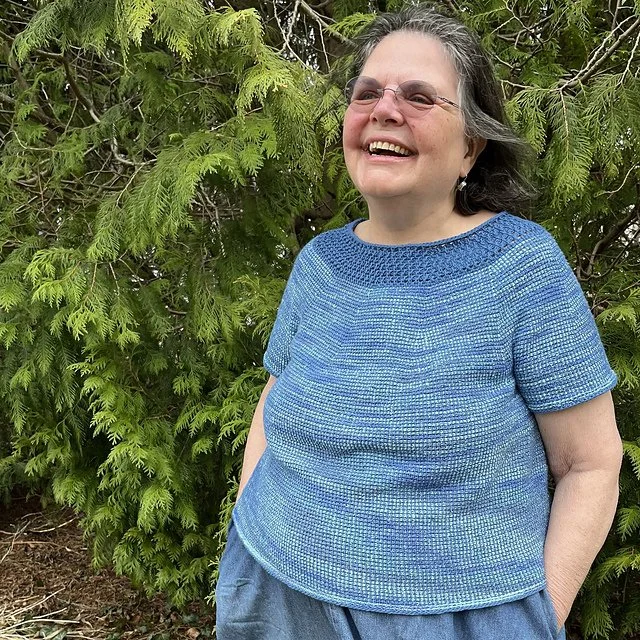 A woman with greying hair and glasses smiling outdoors in front of green foliage.