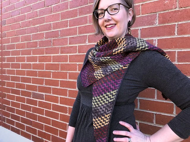 Woman with short gray hair and glasses standing outdoors in front of a red brick wall, smiling, wearing a black dress and a multicoloured patterned shawl.