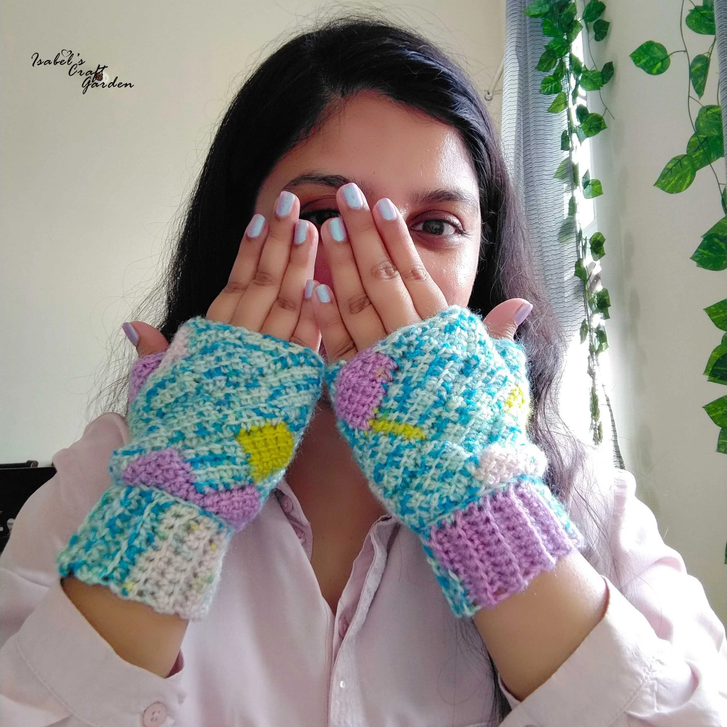 A woman with black hair wearing a white shirt and colourful Tunisian crocheted mittens covering her face partially with her hand, standing near a window with green hanging plants in the background.
