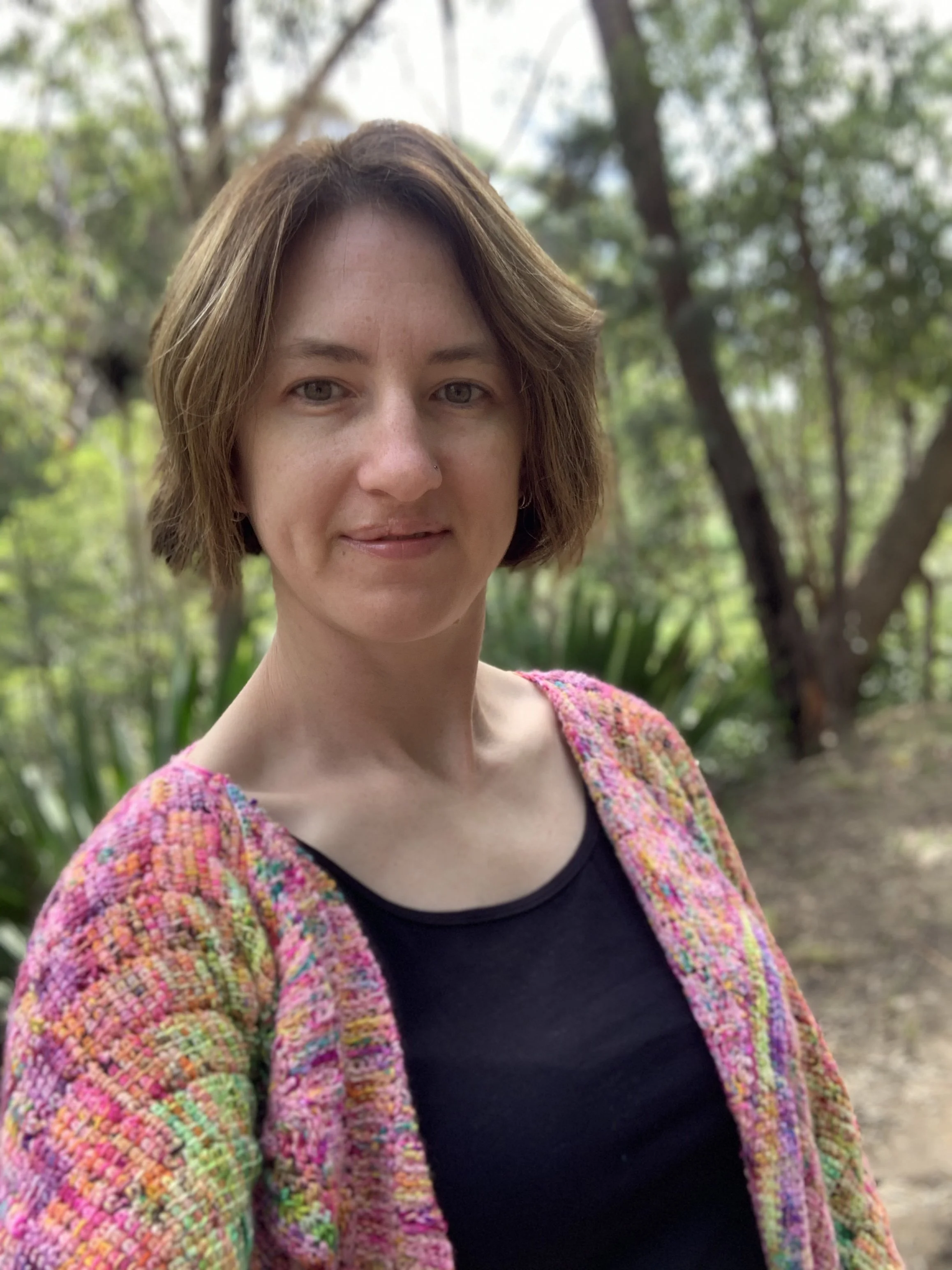 A woman with short brown hair wearing a colourful Tunisian crocheted sweater and a black top, standing outdoors in front of trees and greenery, smiling slightly.