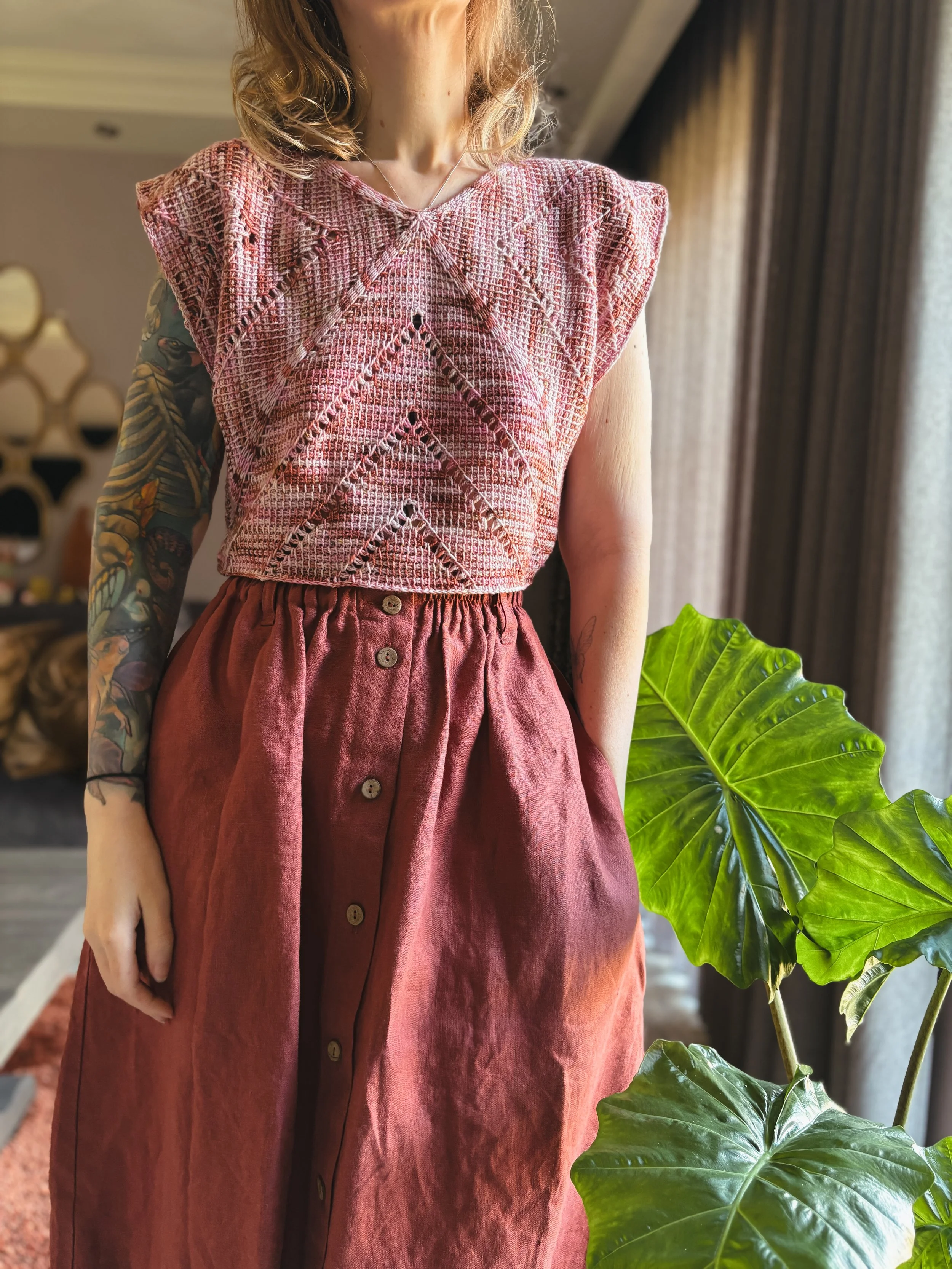 A woman in a pink textured sleeveless top and maroon buttoned skirt standing indoors next to a large green leafy plant.