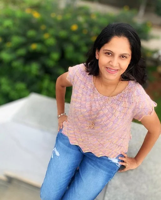 A woman standing outdoors on a sidewalk, smiling, with greenery and yellow flowers in the background. She is wearing a light pink patterned top and blue jeans.