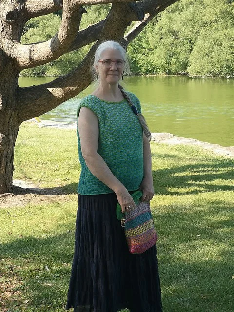A woman with long gray hair, glasses, wearing a green and blue sleeveless top and a long black skirt, standing outdoors near a river with green trees and grass, next to a large tree.