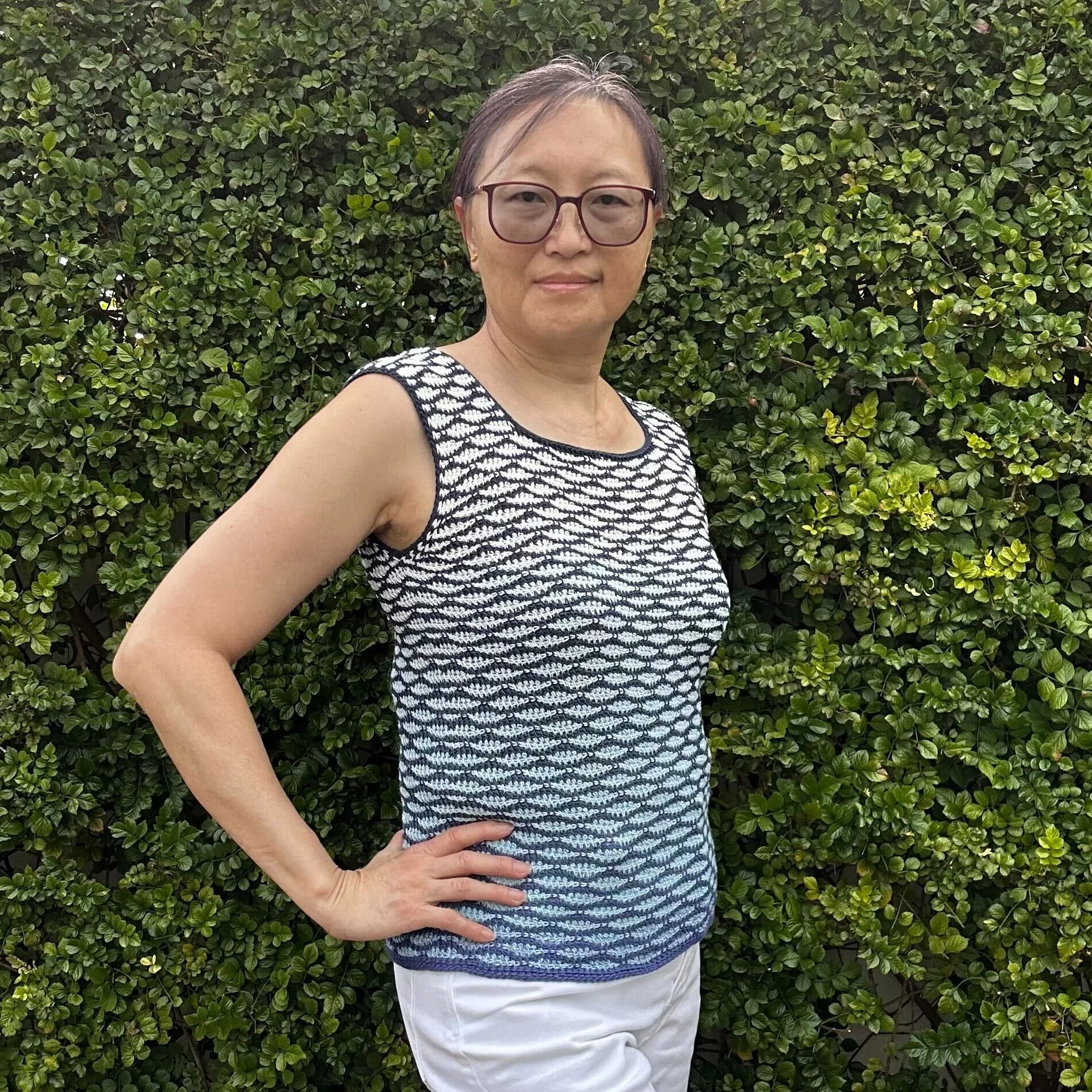 A woman standing outdoors with a hand on her hip, wearing a sleeveless patterned top, glasses, and white pants, in front of a dense green bush.