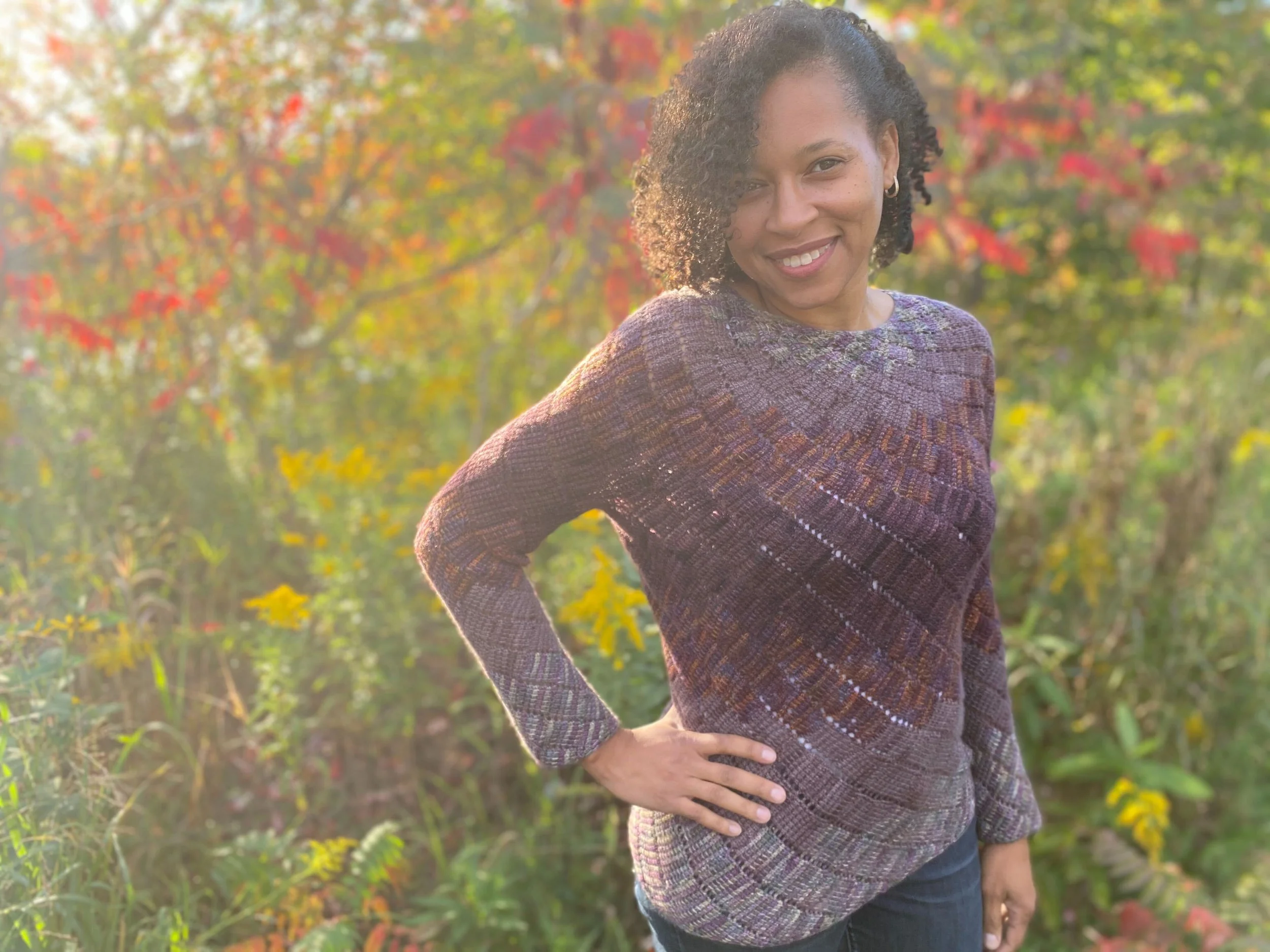 A woman standing outdoors in front of colorful autumn trees, smiling, with one hand on her hip, wearing a multicoloured sweater and jeans.
