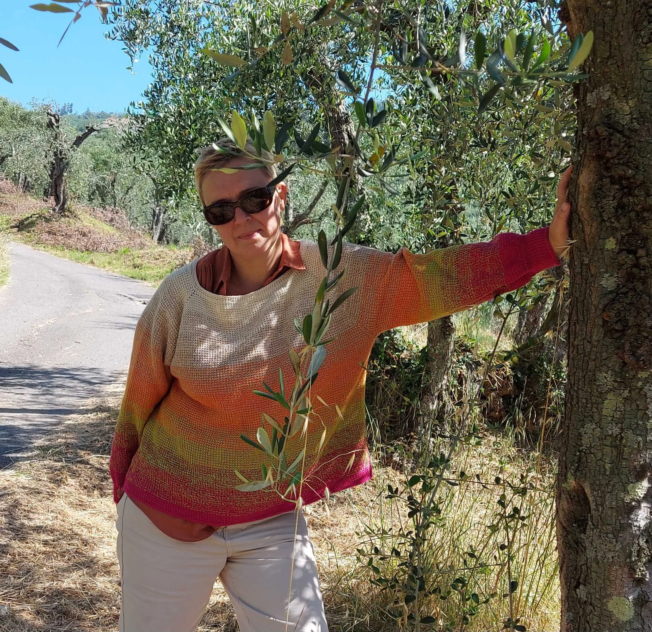 Woman with short blonde hair wearing sunglasses and a multicoloured sweater standing outdoors with her arm around a tree trunk, surrounded by green foliage and bright blue sky.