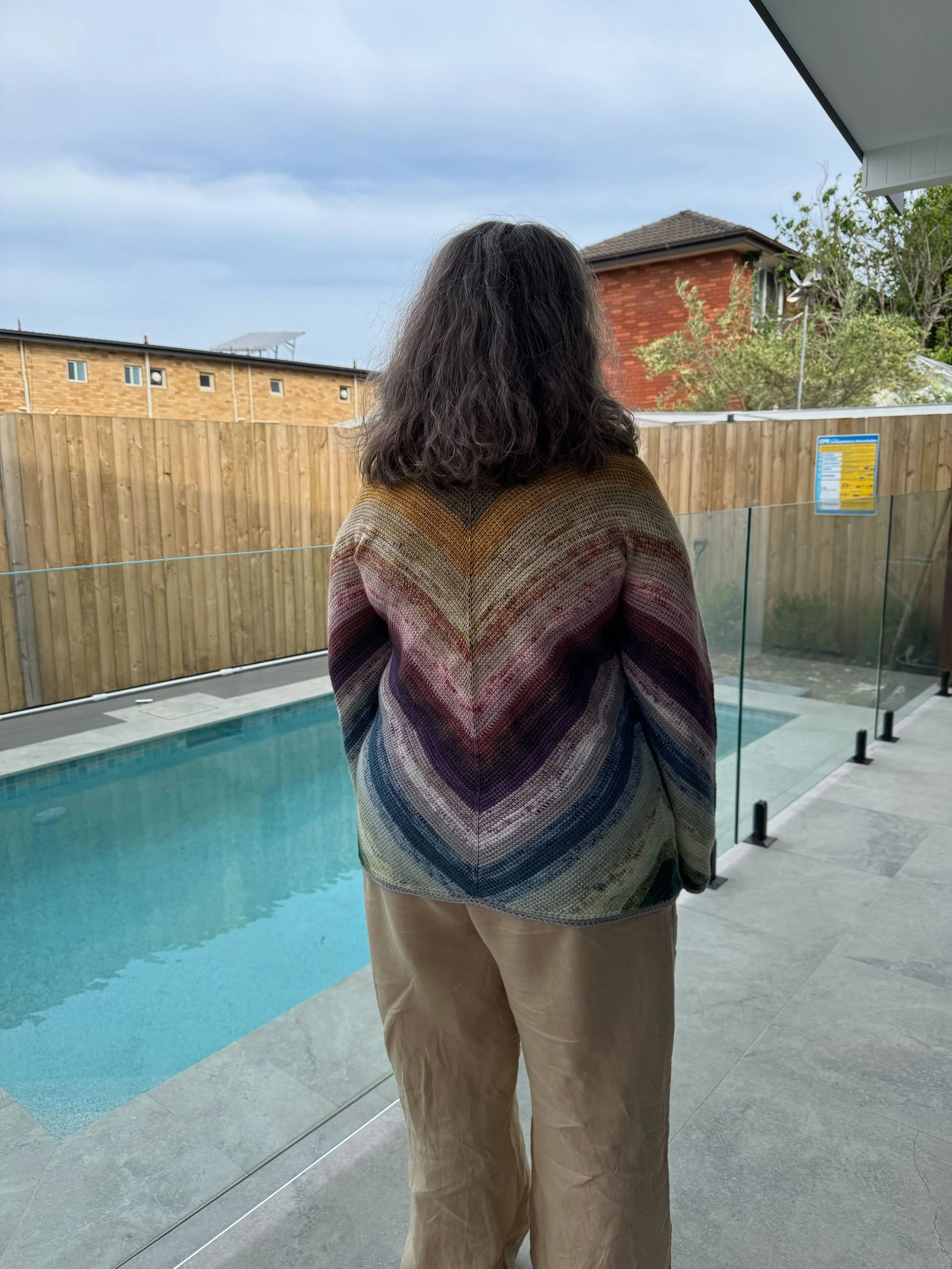 Person with shoulder-length dark hair standing outdoors near a swimming pool, wearing a multicoloured striped sweater and beige pants, looking away from the camera, with a wooden fence, houses, and cloudy sky in the background.