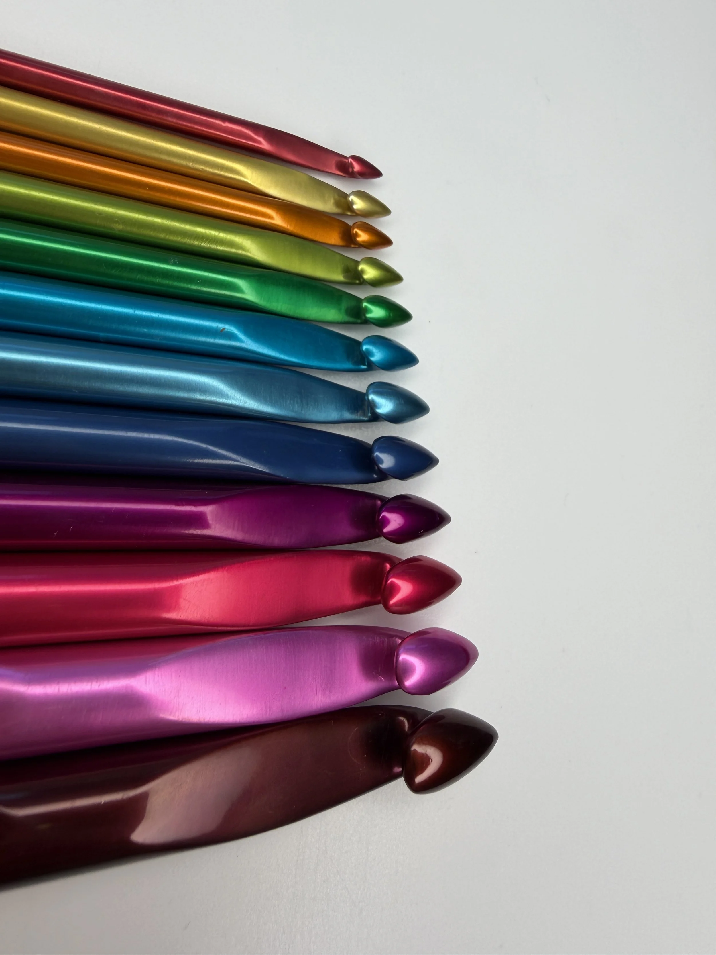 A collection of colourful handmade Tunisian crochet hooks arranged diagonally on a white background, showcasing various colours including red, gold, orange, green, blue, pink, and brown.