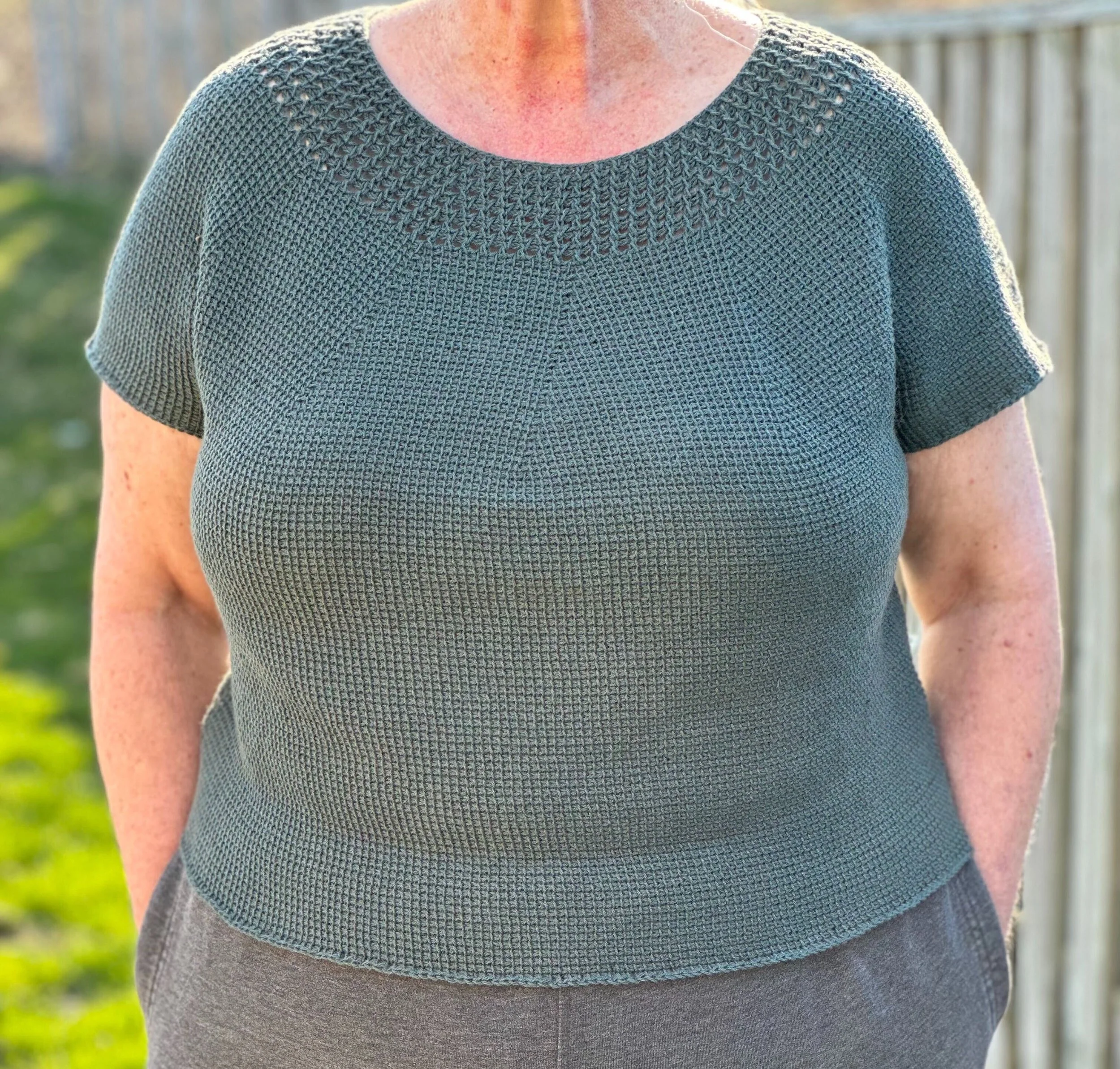 Close-up of a person wearing a green Tunisian crocheted top with a decorative pattern on the shoulders, standing outdoors near a white fence with a blurred green background.