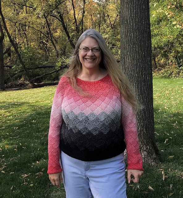 A woman with long gray hair and glasses smiling outdoors next to a tree in a park with green grass and autumn trees in the background.