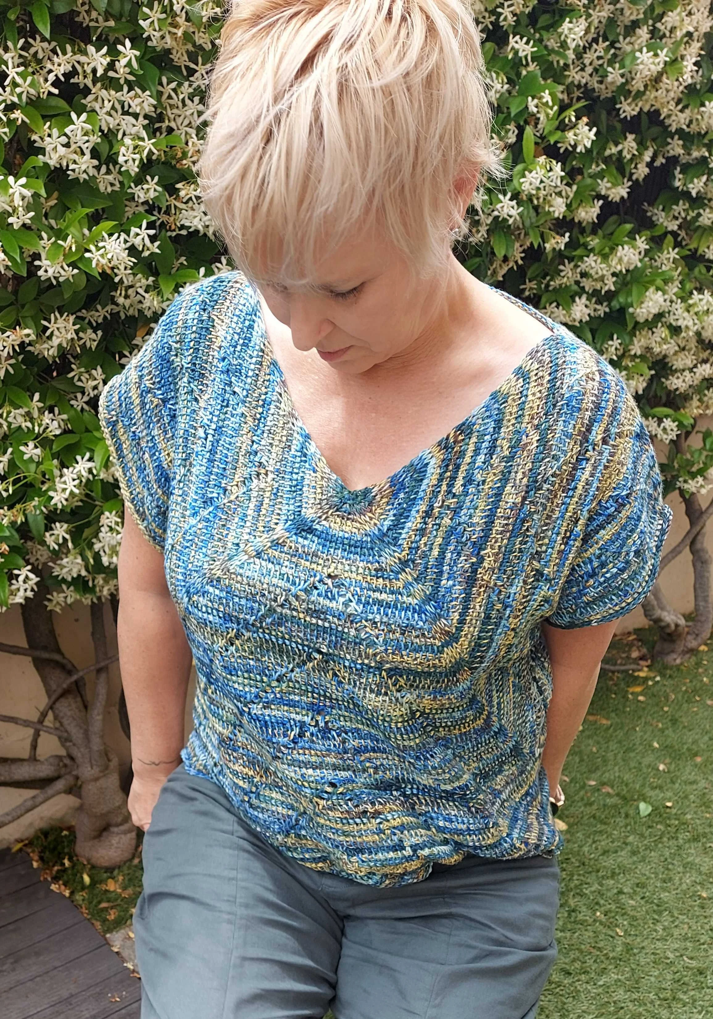 Woman with short blond hair wearing a colourful Tunisian crocheted top and gray pants, sitting outdoors in front of a bush with white flowers.