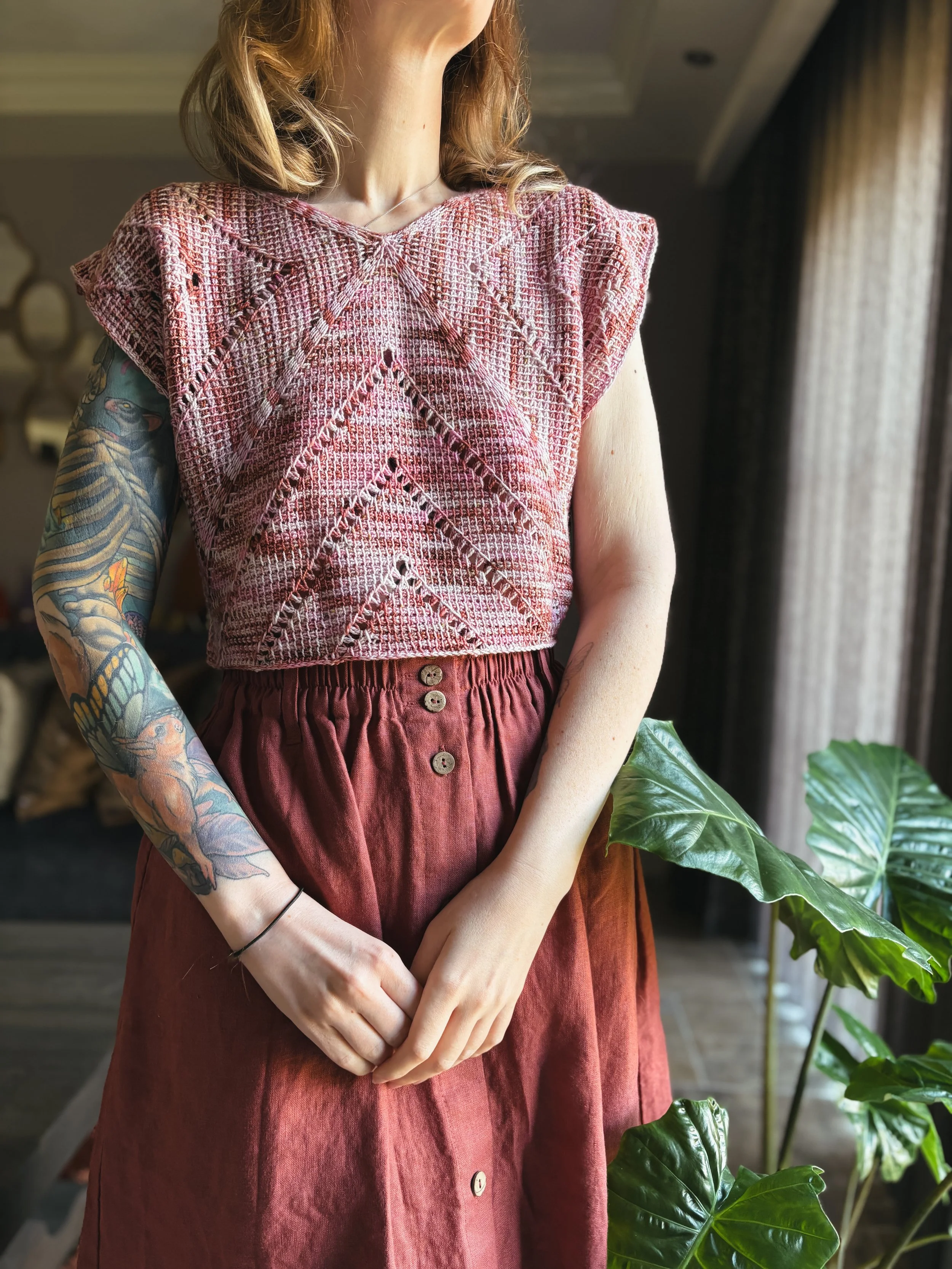 A woman with a tattooed left arm, wearing a pink patterned sleeveless top and red high-waisted skirt, standing next to green plants in a room with curtains and a window.