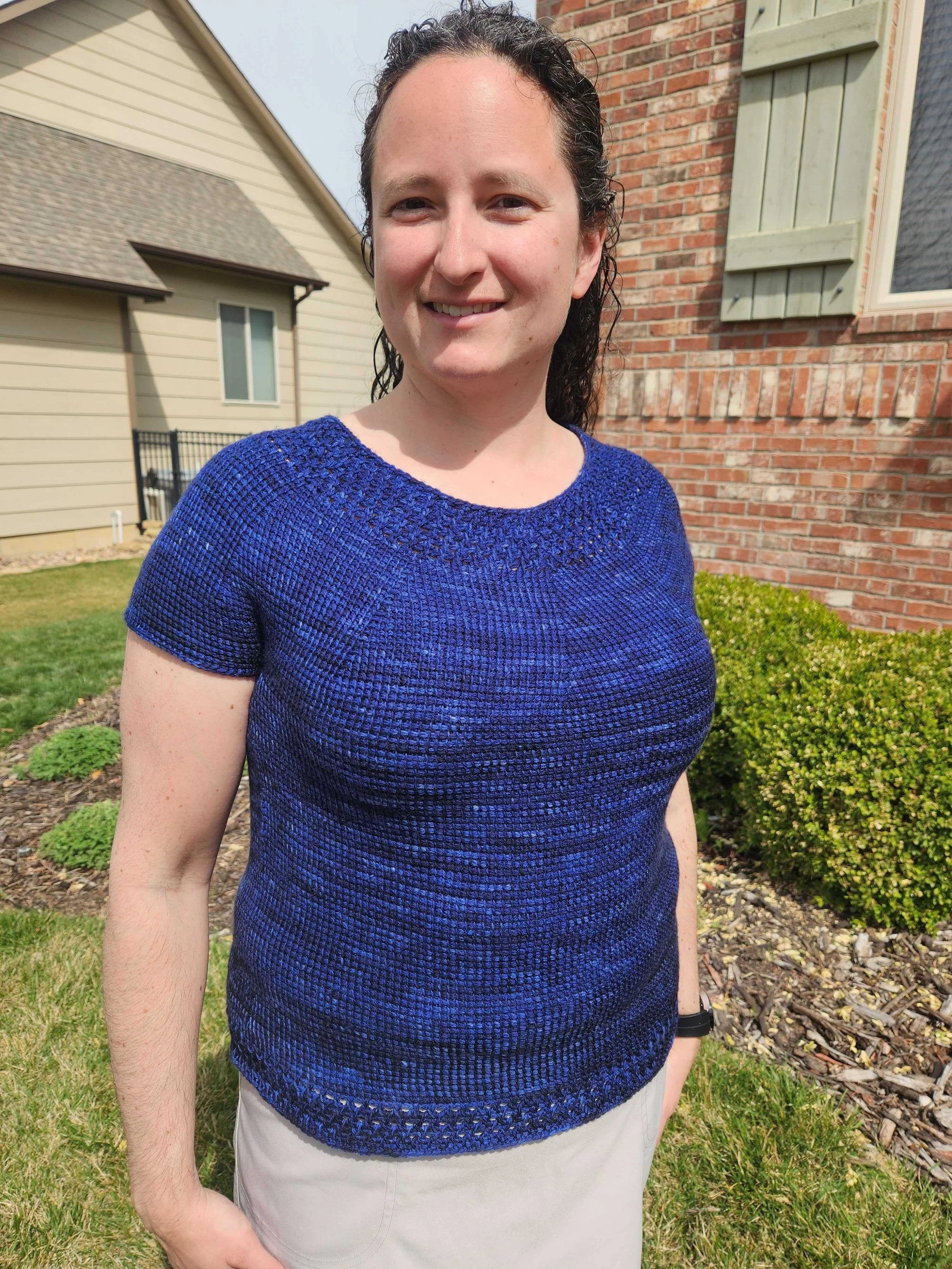 A woman with curly dark hair smiling outdoors, wearing a blue Tunisian crocheted top and beige pants, standing in front of a brick house with a window and green bushes.