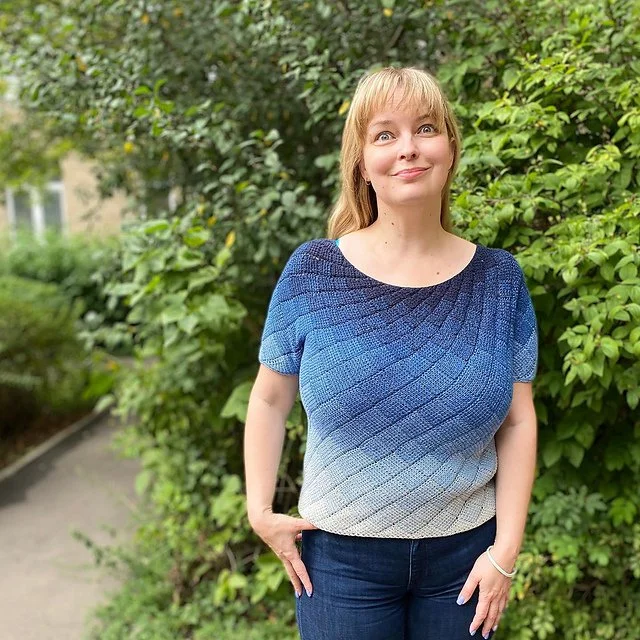 A woman with blonde hair and a blue gradient top stands outdoors in front of green bushes, smiling softly.