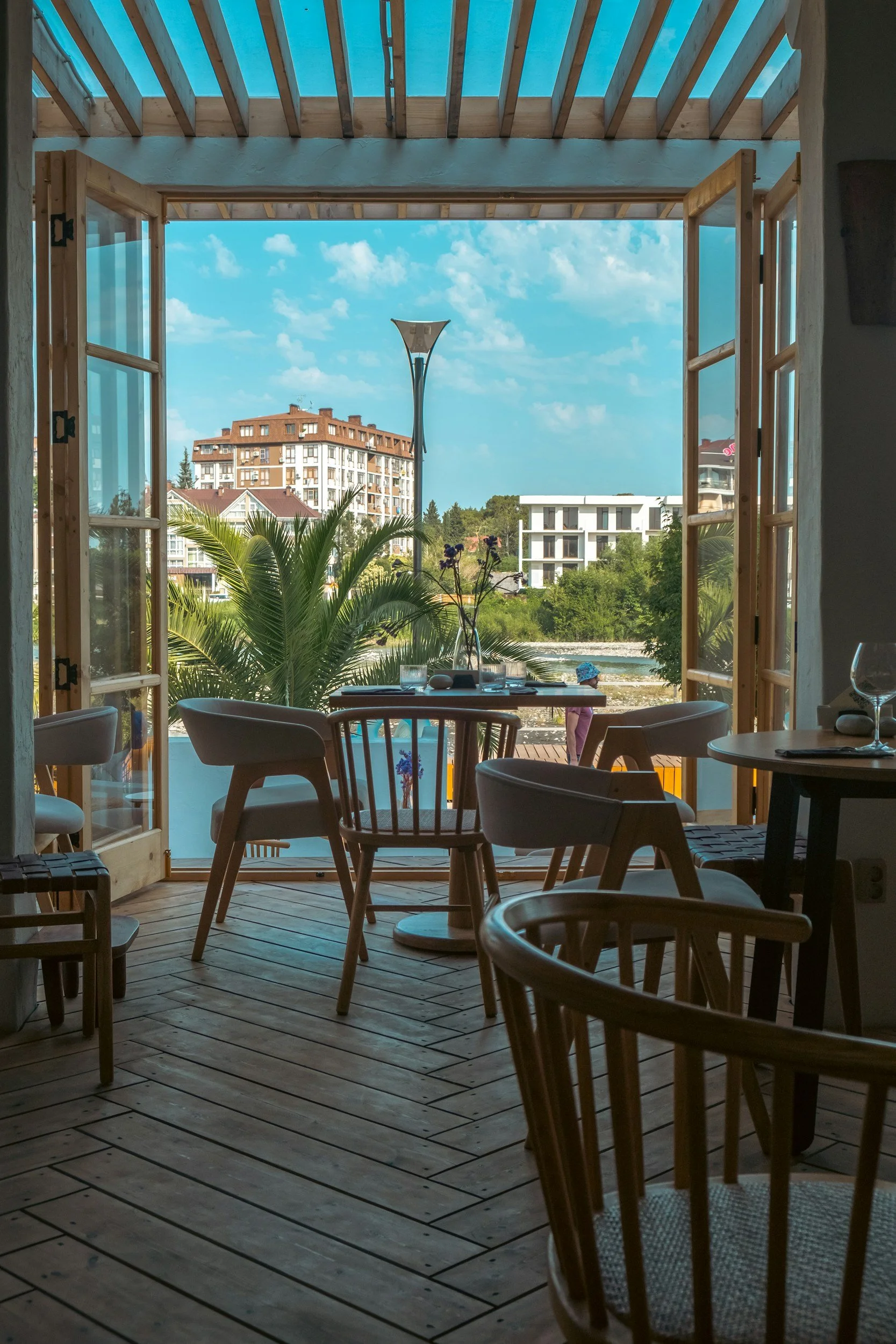 View from inside a café or restaurant with wooden flooring, empty chairs, and large open windows revealing a sunny outdoor scene with palm trees, a street lamp, a woman with a hat, and several buildings against a blue sky with some clouds.