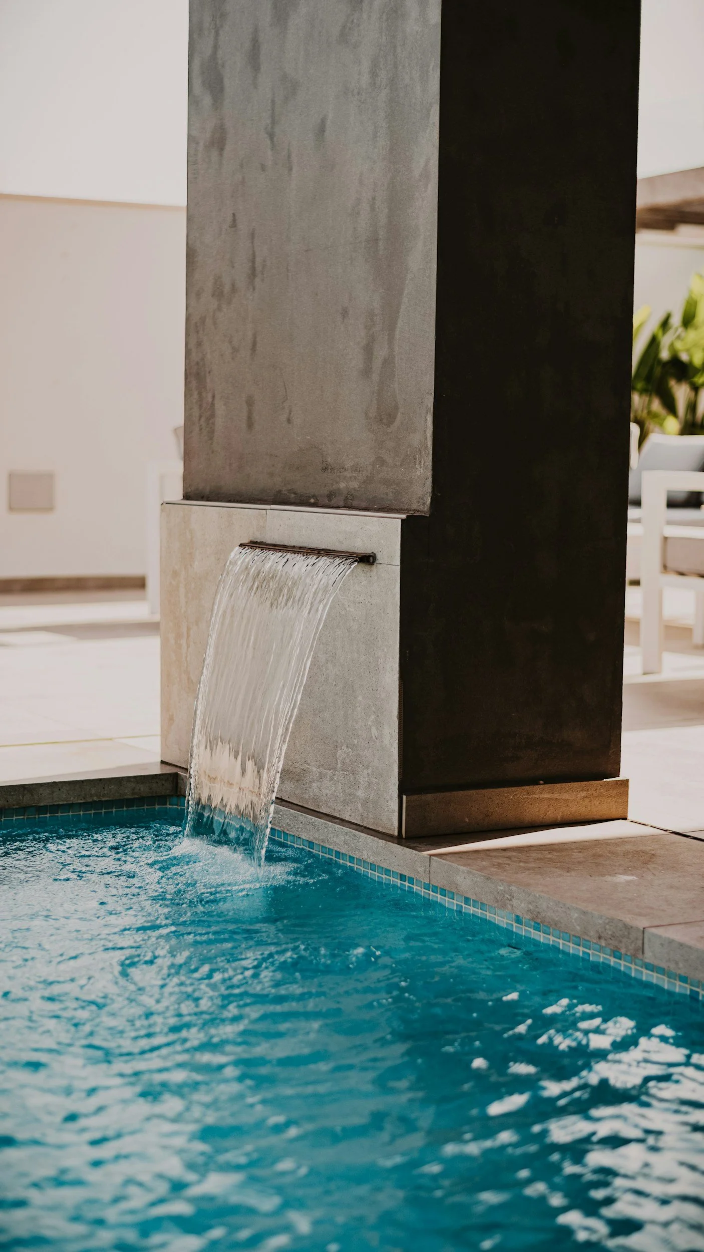A modern outdoor water feature with water cascading from a spout into a swimming pool, surrounded by a beige tile deck and dark vertical panels.