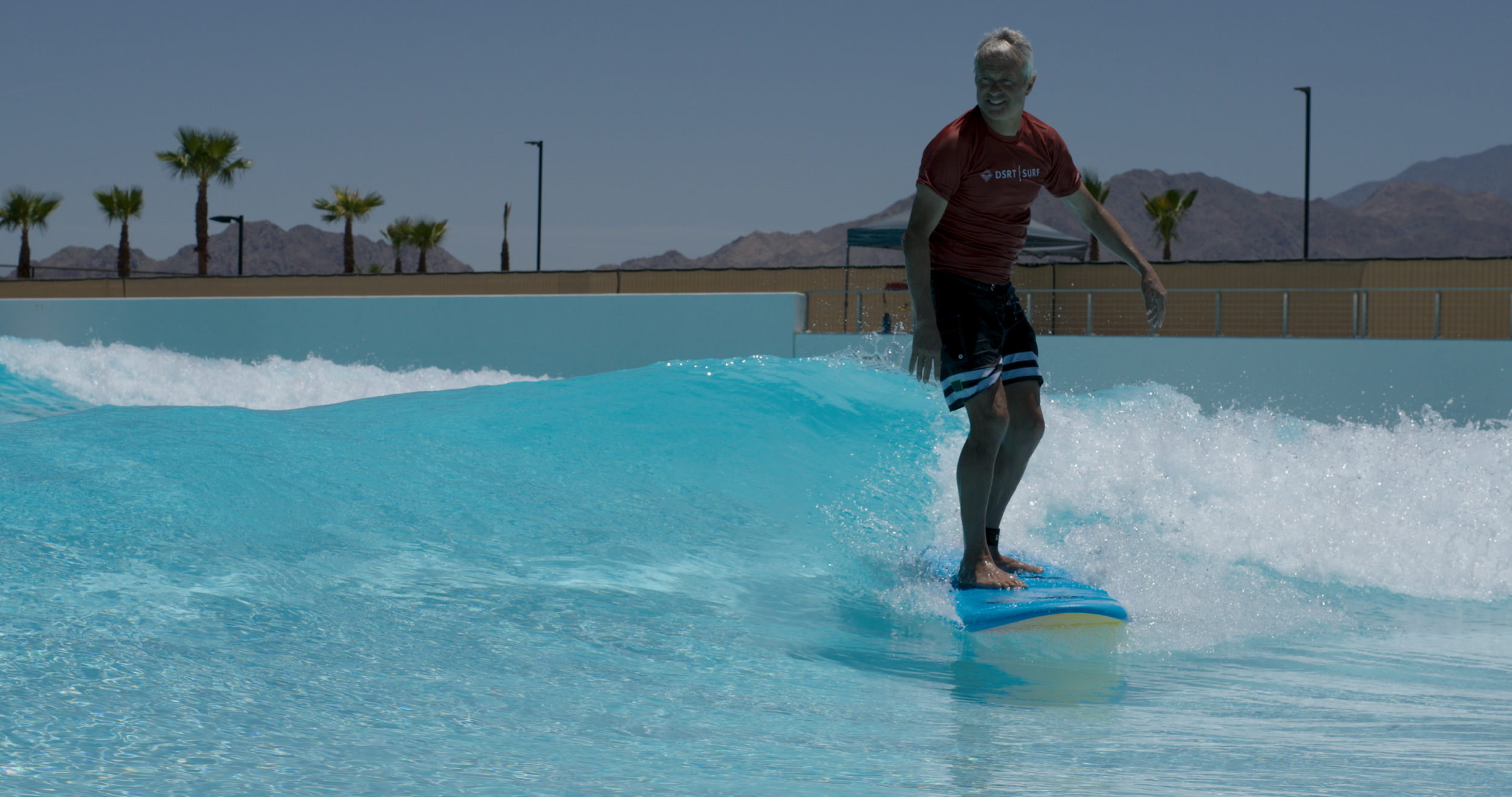 A man surfing on a wave at a water park or pool, with palm trees and mountains in the background on a sunny day.