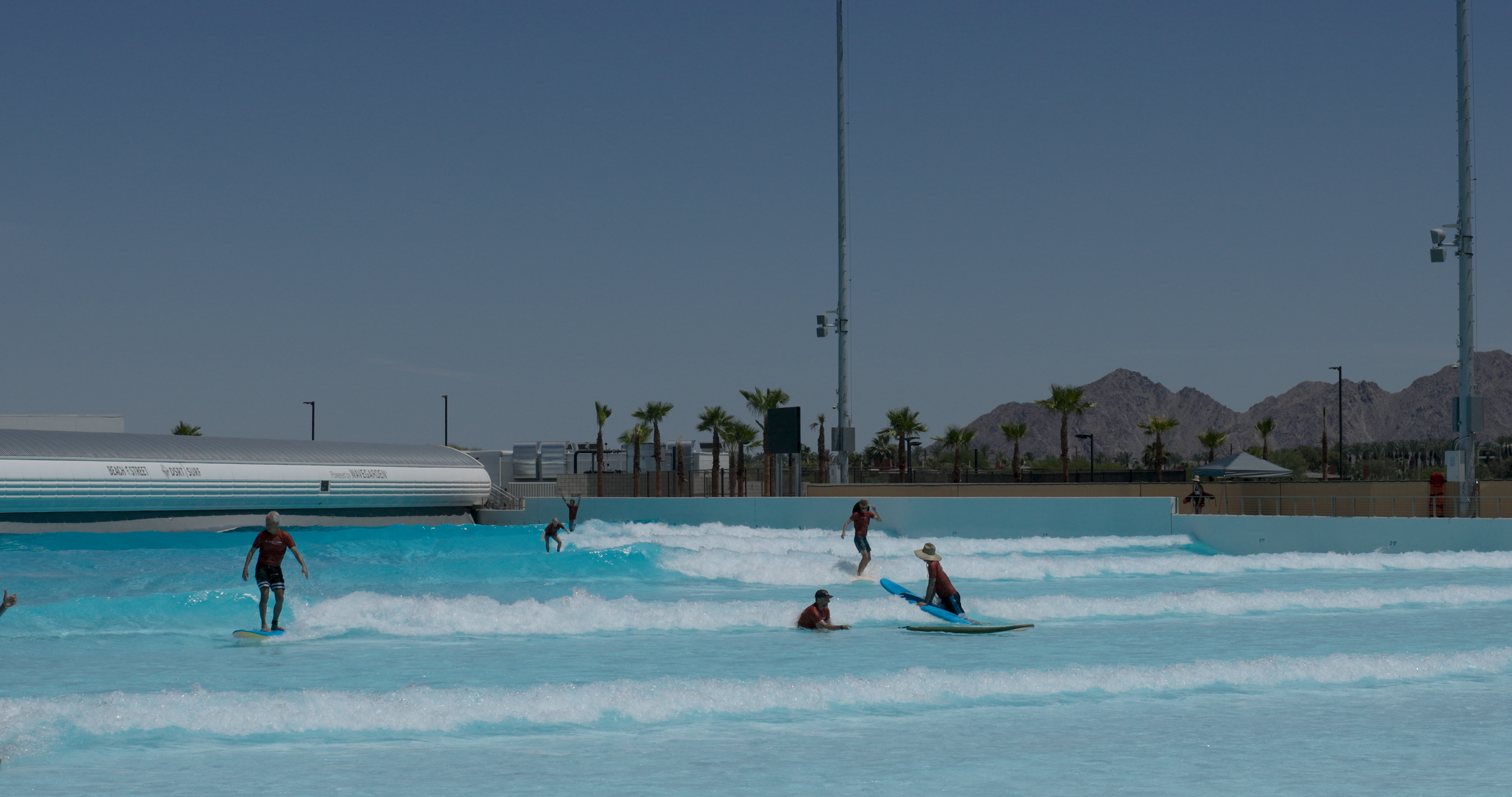 People surfing and enjoying a wave pool with palm trees, mountains, and blue sky in the background.