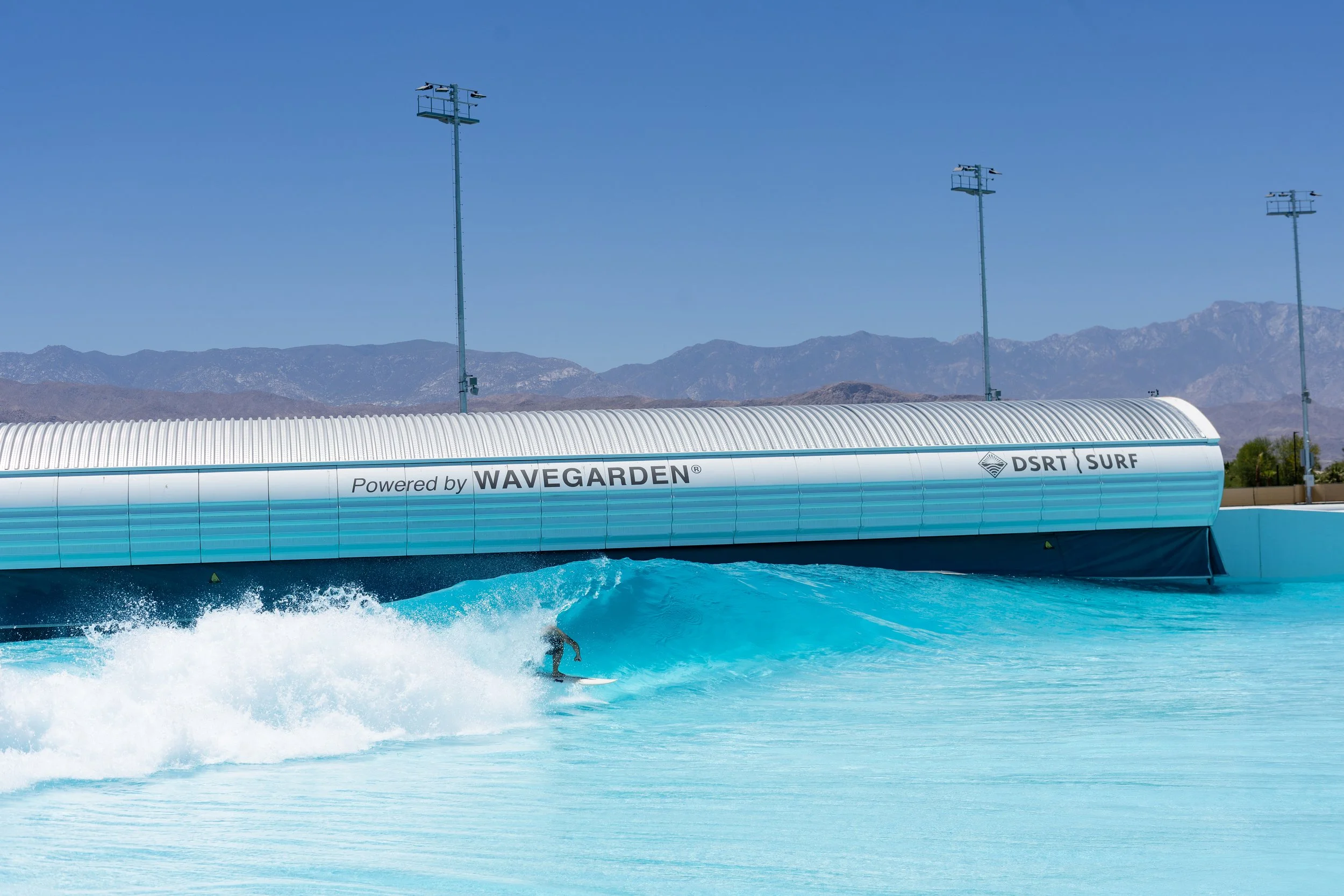 A water ride at an amusement park with a large splash in the pool and a person surfing the wave, with the ride's tunnels and control towers labeled 'Powered by Wavegarden' and 'DSRT Surf' in the background, and mountains under a clear blue sky.