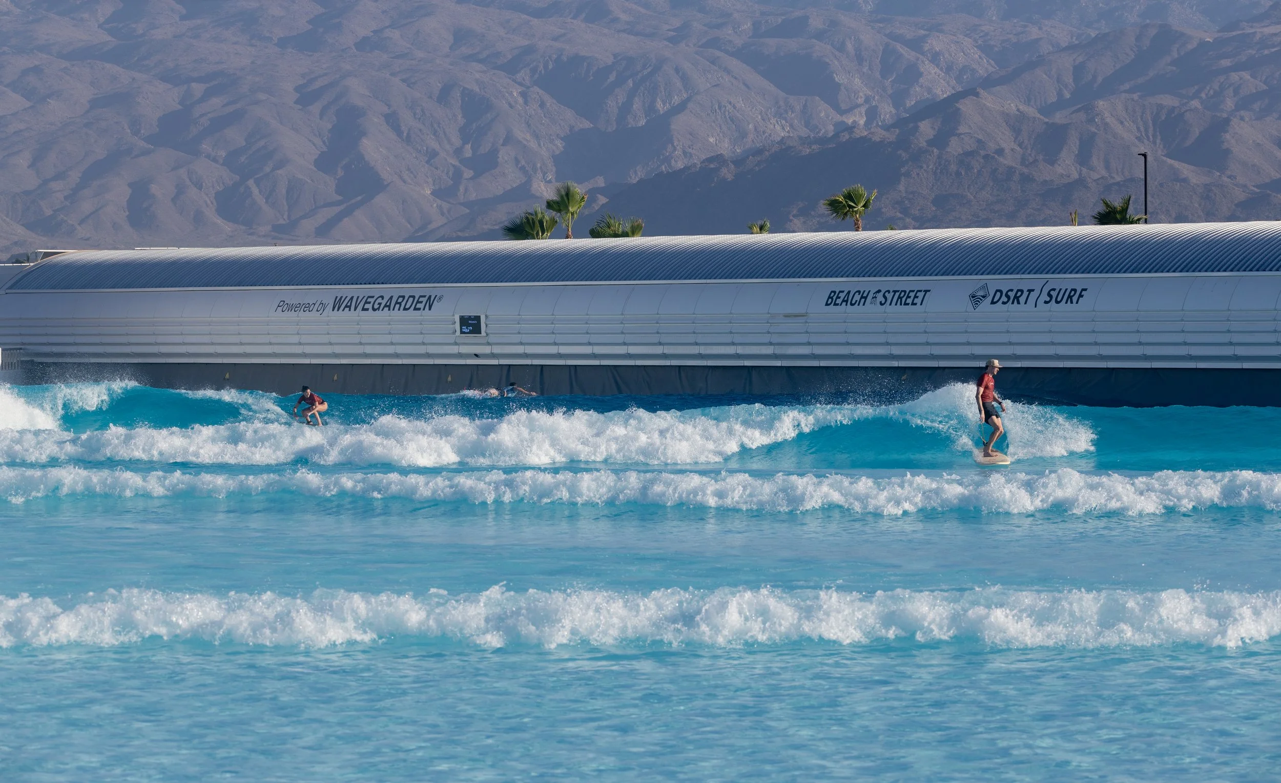 People surfing in a wave at an artificial surfing facility with a modern structure and mountainous background.