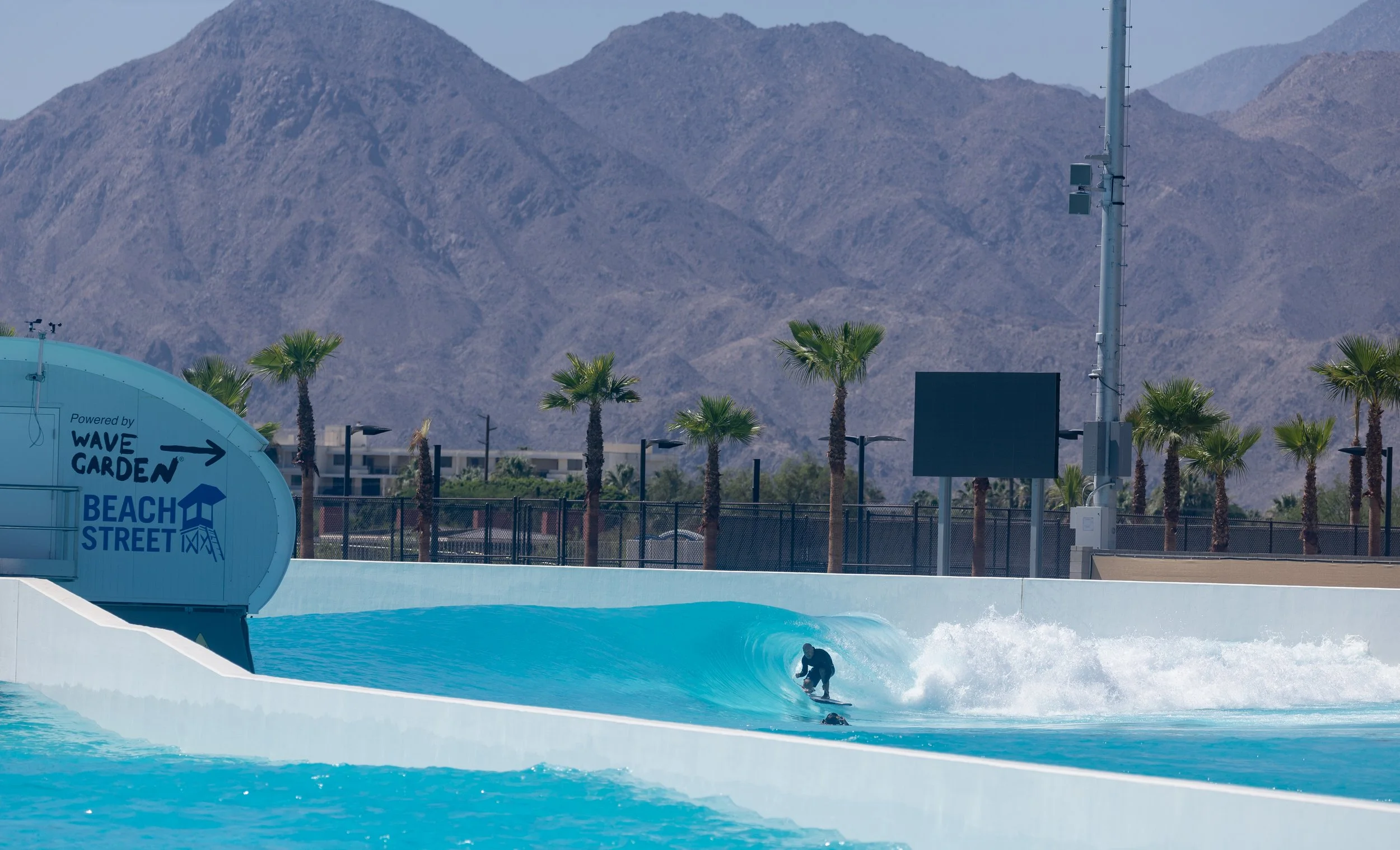 A person surfing on a wave at a water park with a backdrop of mountains, palm trees, and a clear sky.
