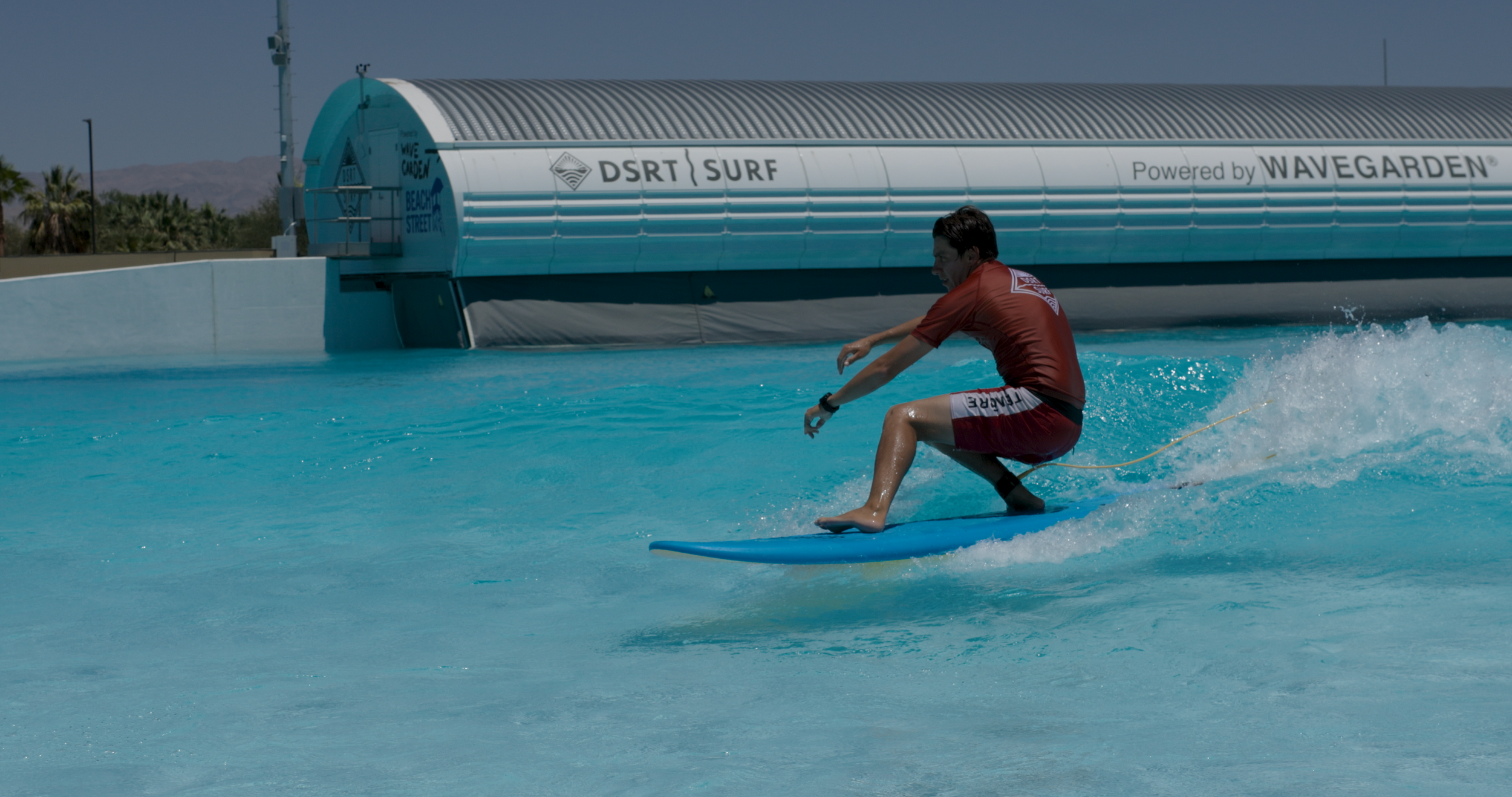 A person surfing on a wave at a water park with a large water slide and palm trees in the background.
