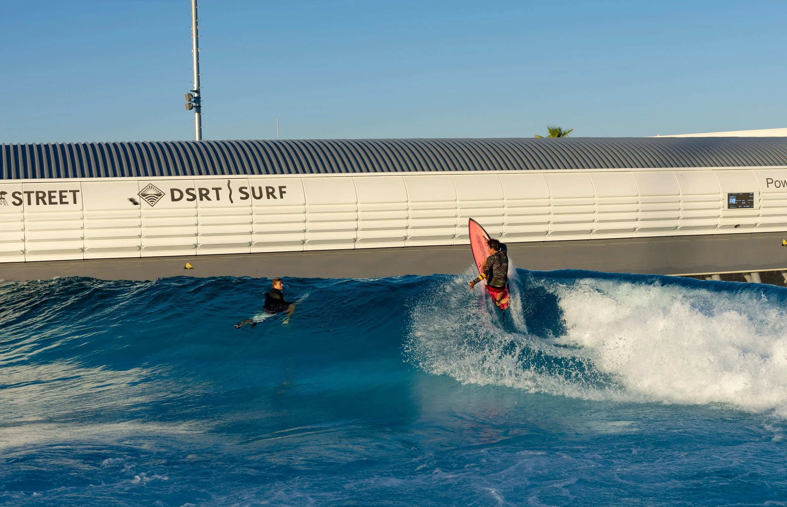 A person riding a wave on a surfboard with another person in the water nearby, and a large industrial or commercial building in the background.