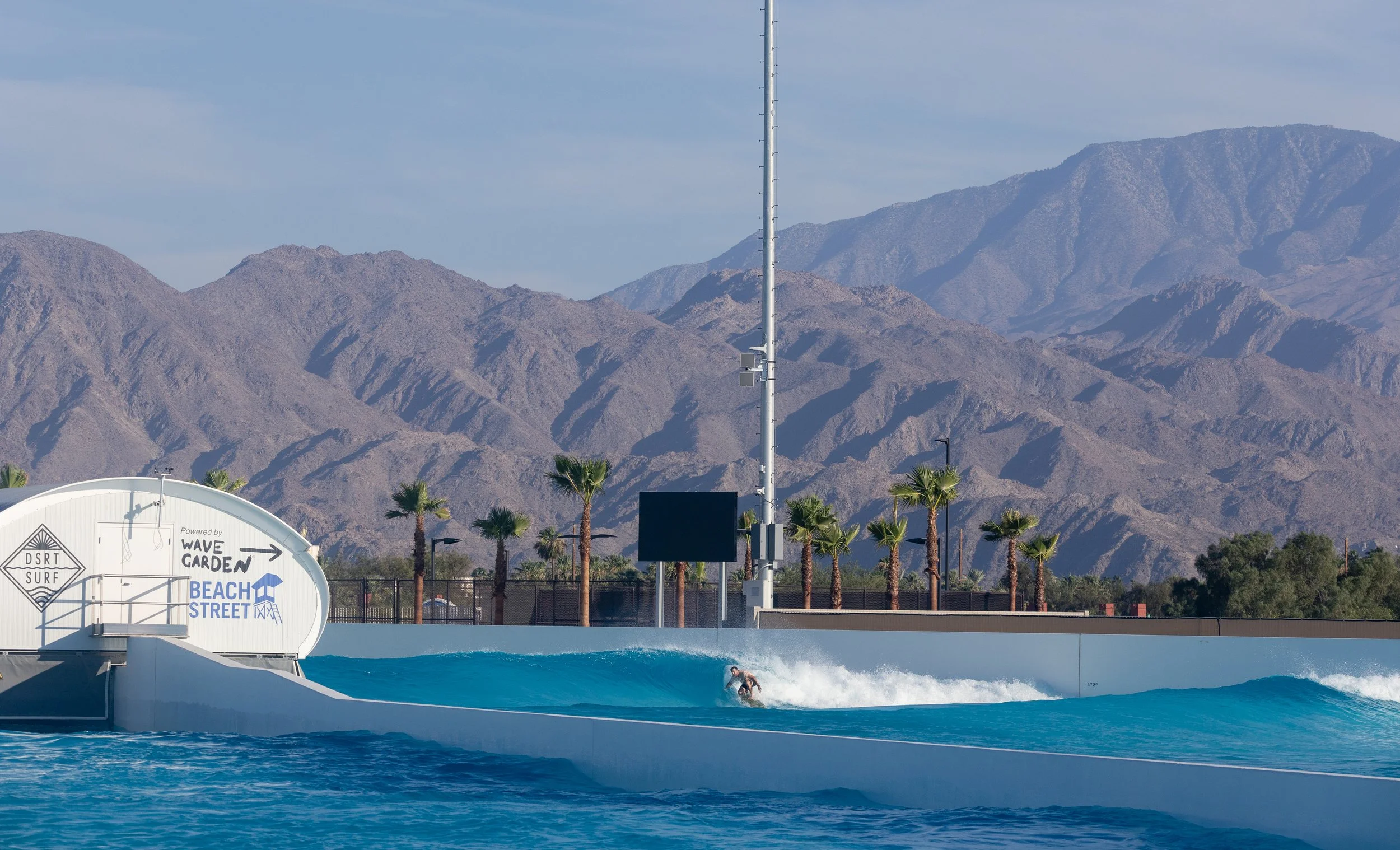 A surfer riding a wave at a man-made surf park with a backdrop of mountains, palm trees, and a sign that reads 'Wave Garden Beach Street'.