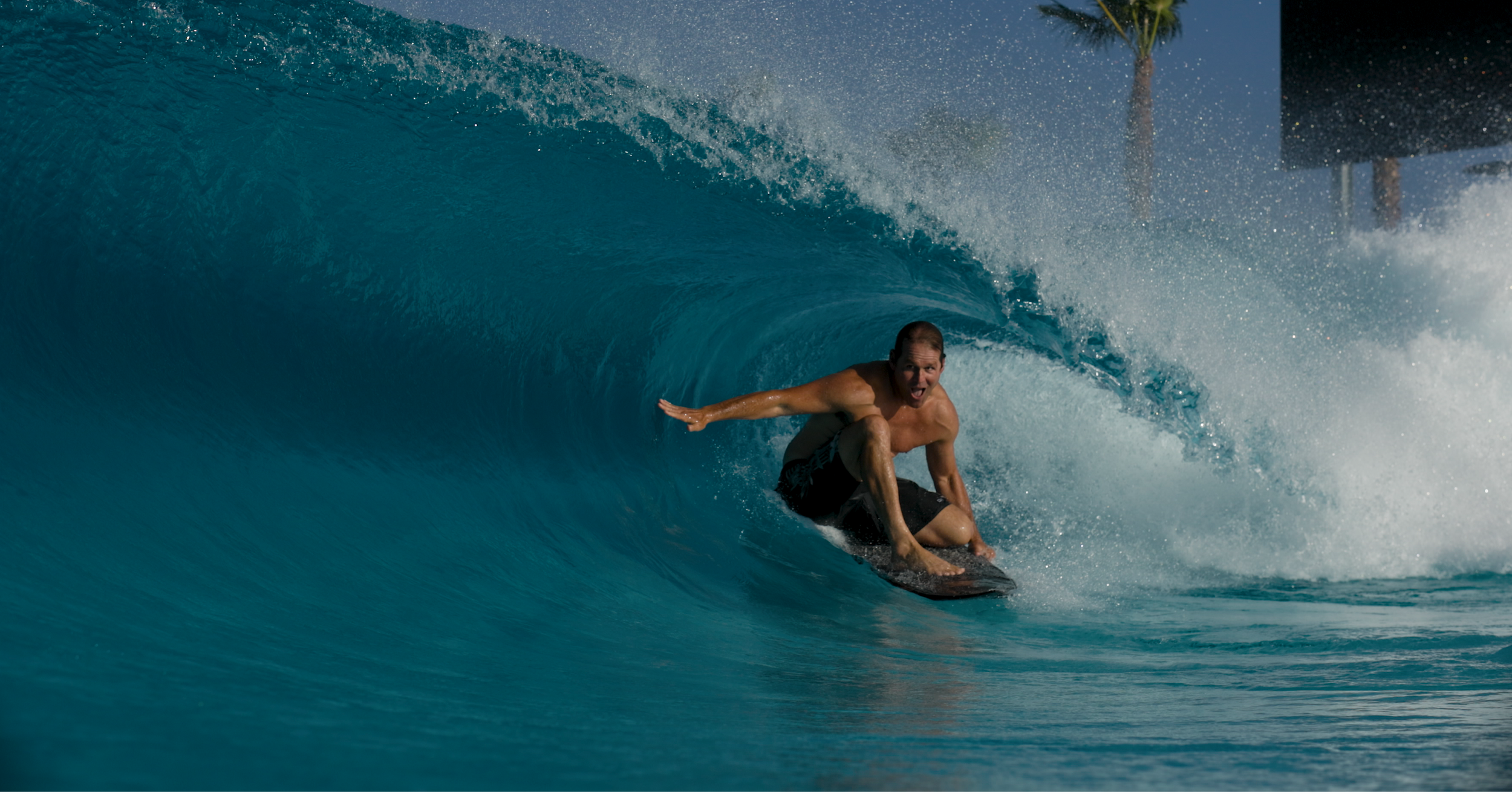 People surfing on artificial wave at a wave pool with mountain landscape in the background.