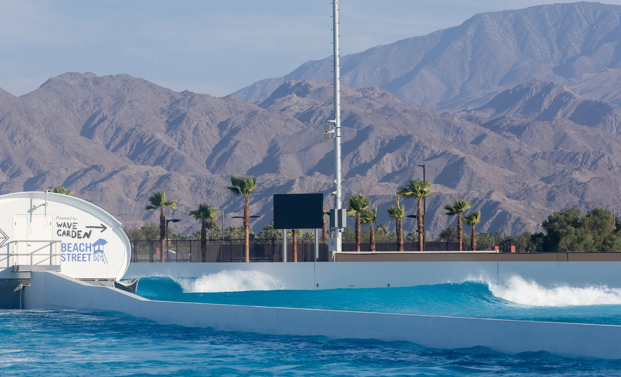 Water park with a wave pool, palm trees, a sign indicating 'Beach Street,' and a mountain range in the background.