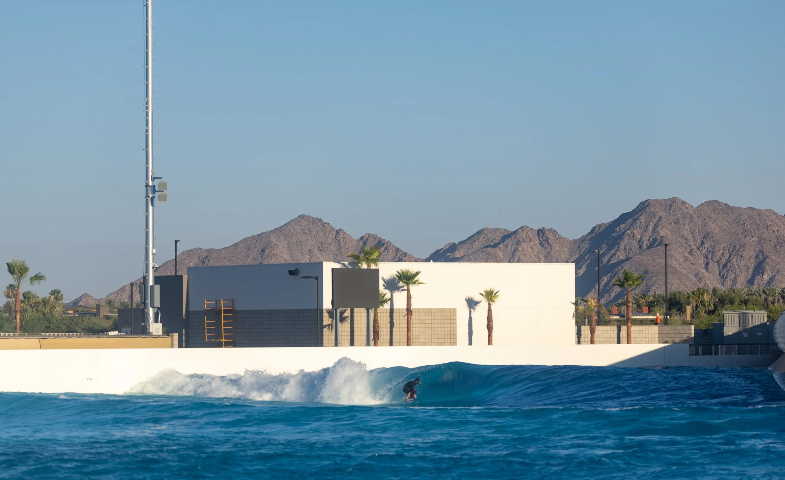 A surfer riding a wave with a white building and palm trees in front of distant mountains under a clear blue sky.