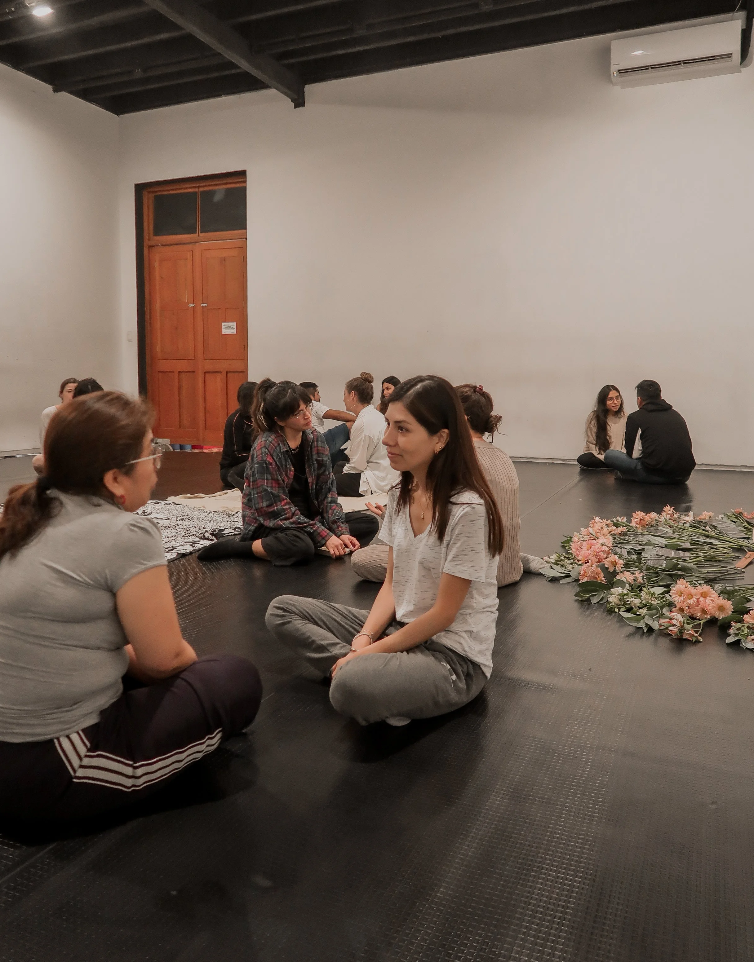 Grupo de personas sentadas en el suelo, en un entorno de calma y bienestar, formando círculos en un espacio interior con paredes blancas y una puerta de madera al fondo, algunas con flores sobre la alfombra.