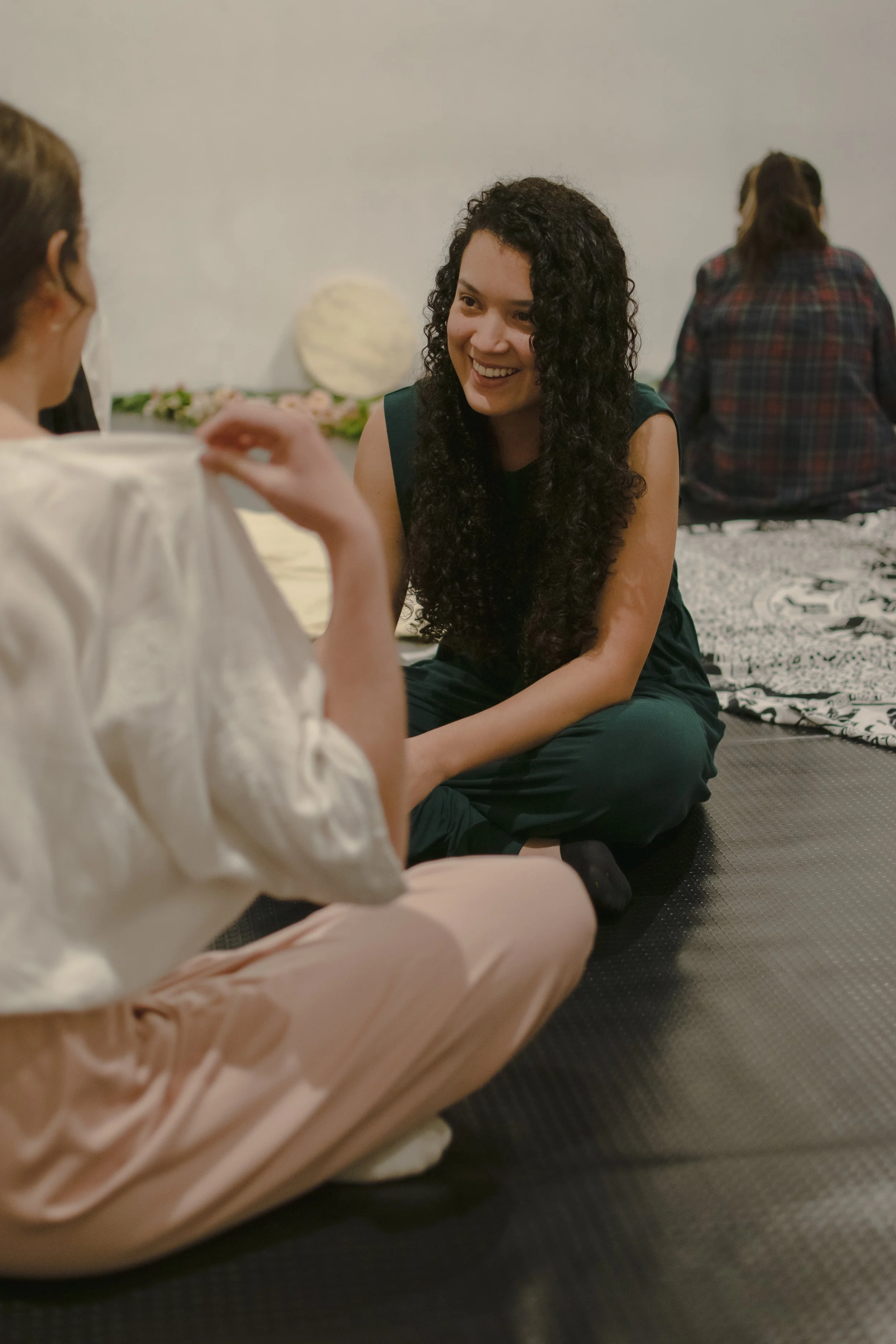 Dos mujeres conversando, una de ellas riendo, en un espacio de bienestar, promoviendo comunicación consciente y conexión en un ambiente relajado. Al fondo, otra persona sentada con la espalda volteada.