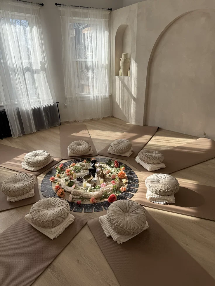 Sala de yoga con almohadillas y cojines en círculo, adornada con flores y estatua en el centro, con luz natural entrando por las cortinas, ideal para talleres, retiros y experiencias de bienestar para personas y organizaciones.