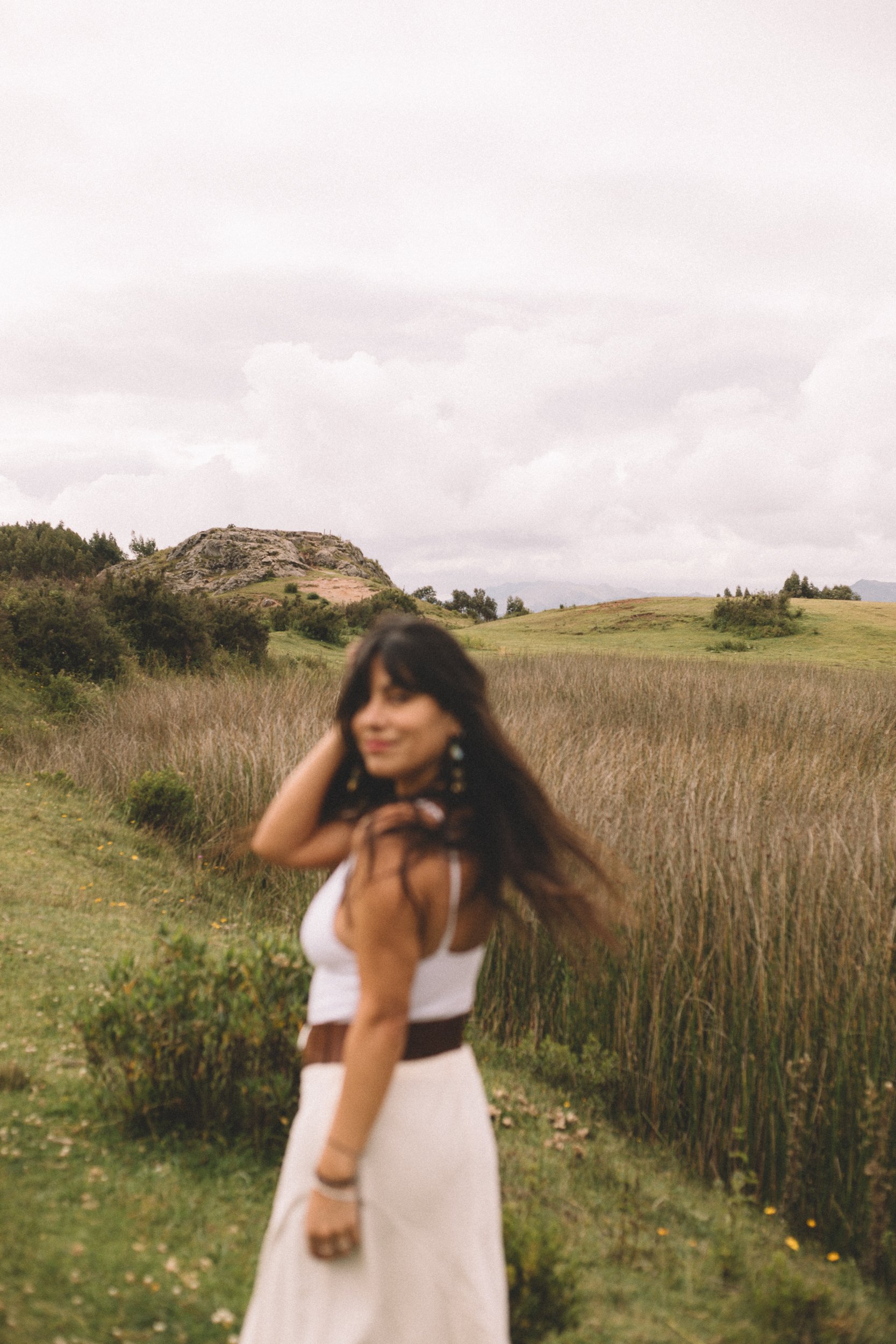 Mujer con vestido largo en un campo de hierba con colinas y una roca grande al fondo, cielo nublado, invitando a escribir o consultar para experiencias personalizadas.