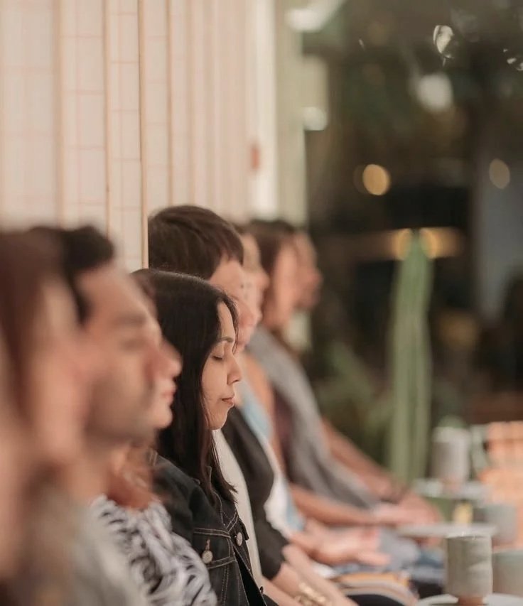 Varios jóvenes sentados en una fila, meditando con los ojos cerrados, en un ambiente de calma con iluminación suave.