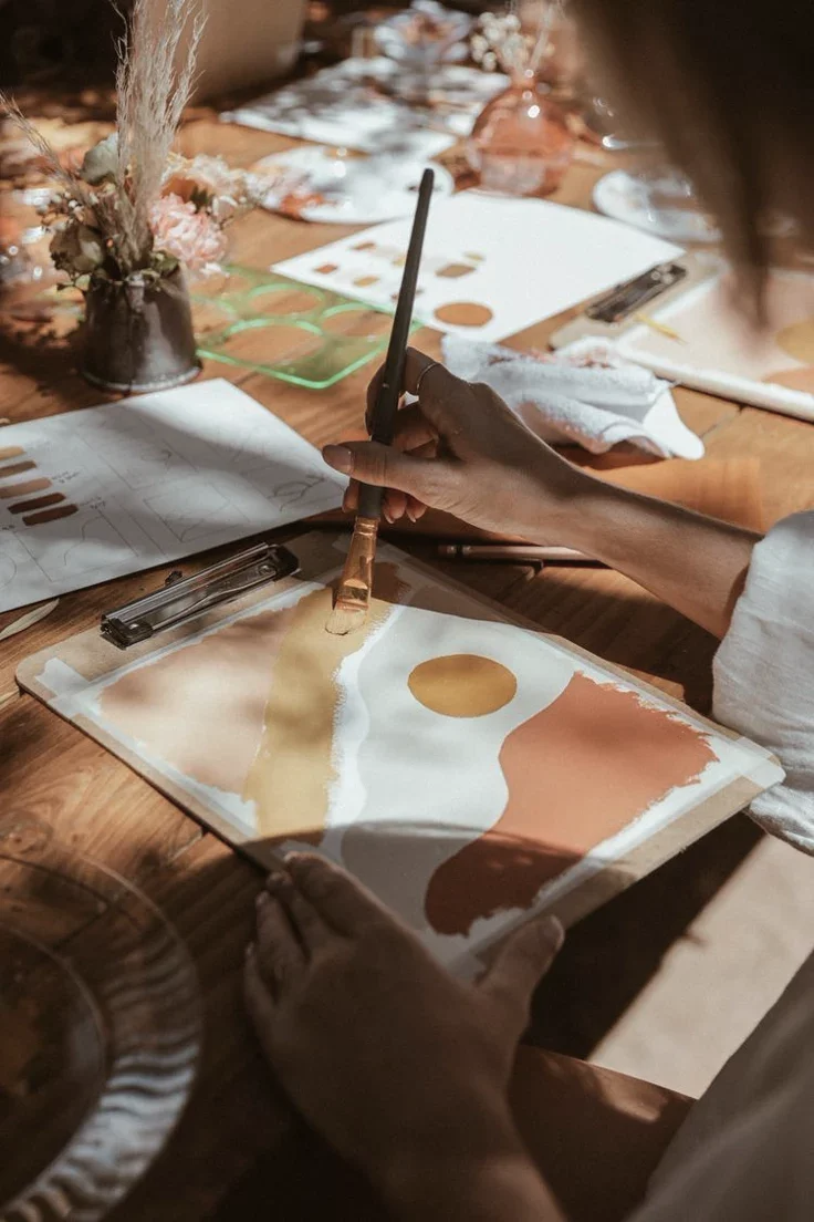 Persona haciendo trabajo creativo, pintando con acuarelas en una mesa de madera, con papeles y pinceles, en un ambiente luminoso y decorado con flores.