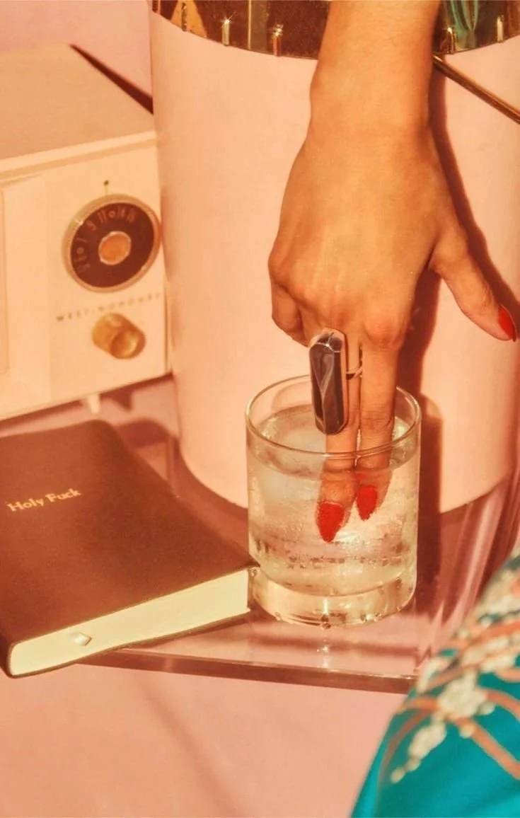 Mano con uñas rojas y anillo en el dedo medio, sumergiendo el dedo medio en un vaso con agua.