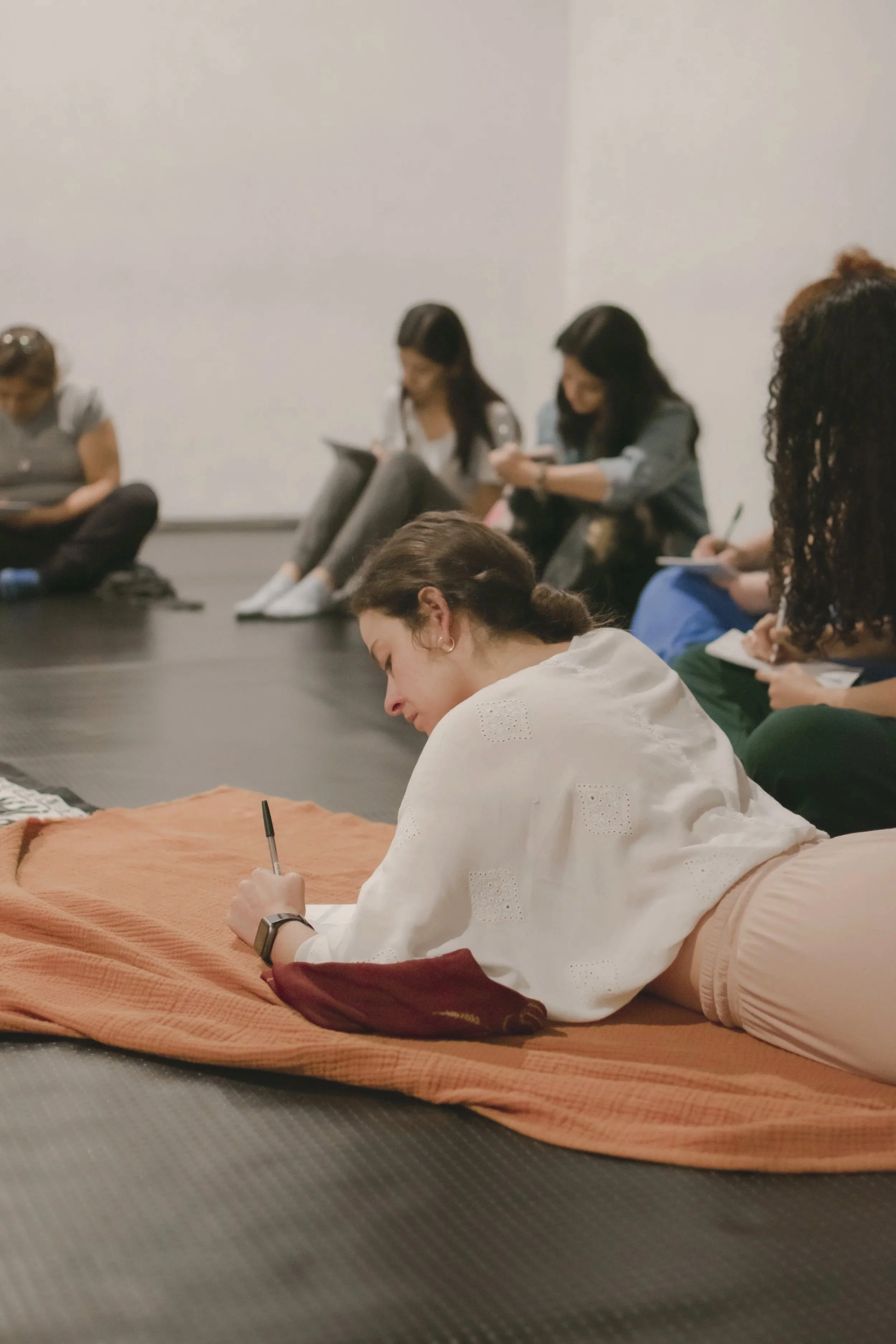 Personas tomando notas en una clase o taller, algunas sentadas en el suelo y otras en posición de descanso, enfocadas en sus cuadernos o dispositivos electrónicos en un ambiente relajado.