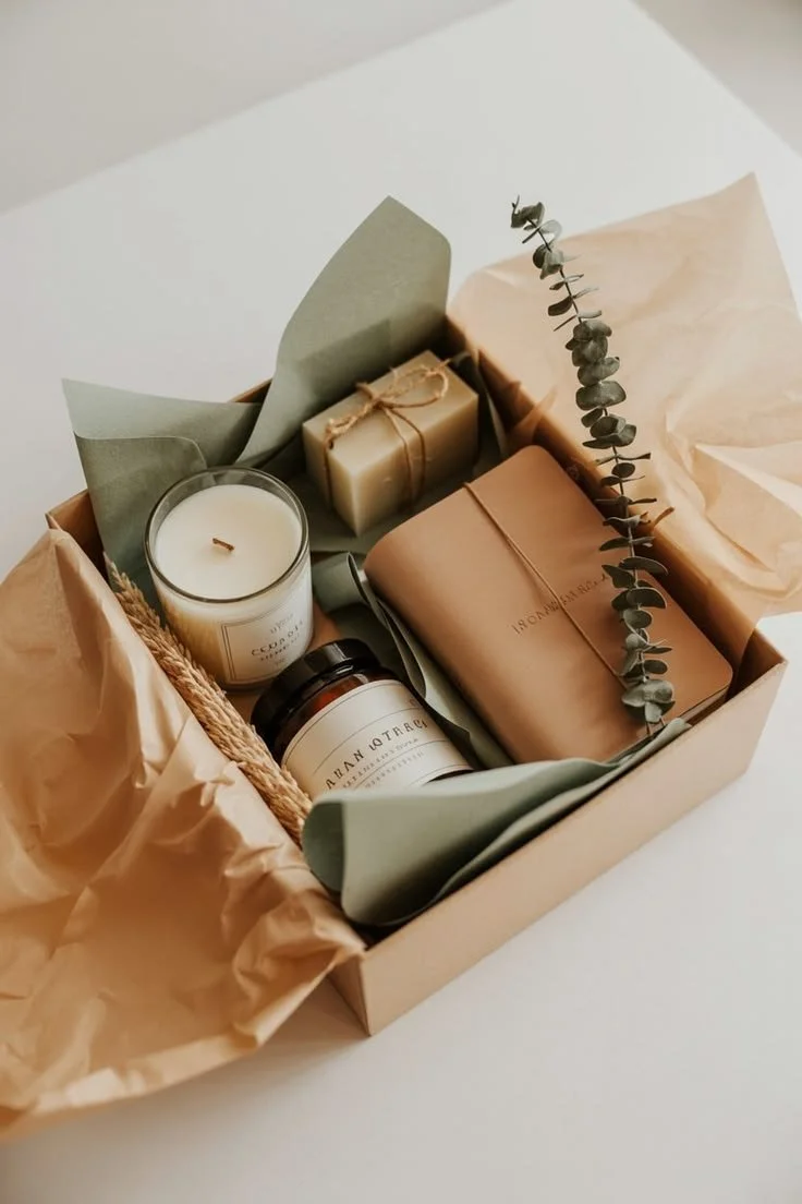 kit de bienestar con velas, jabón, cuaderno y adorno de eucalipto y trigo, envueltos en papel ecológico de colores neutros.