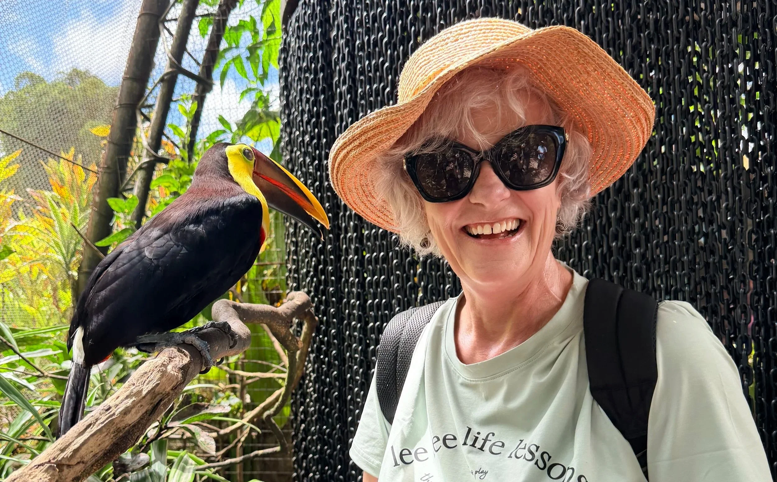 An older woman wearing a large straw sunhat, dark sunglasses, and a light-colored t-shirt with text, smiling while taking a selfie with a colorful toucan perched on a branch beside her. The background includes green foliage and a black chain-link fence.