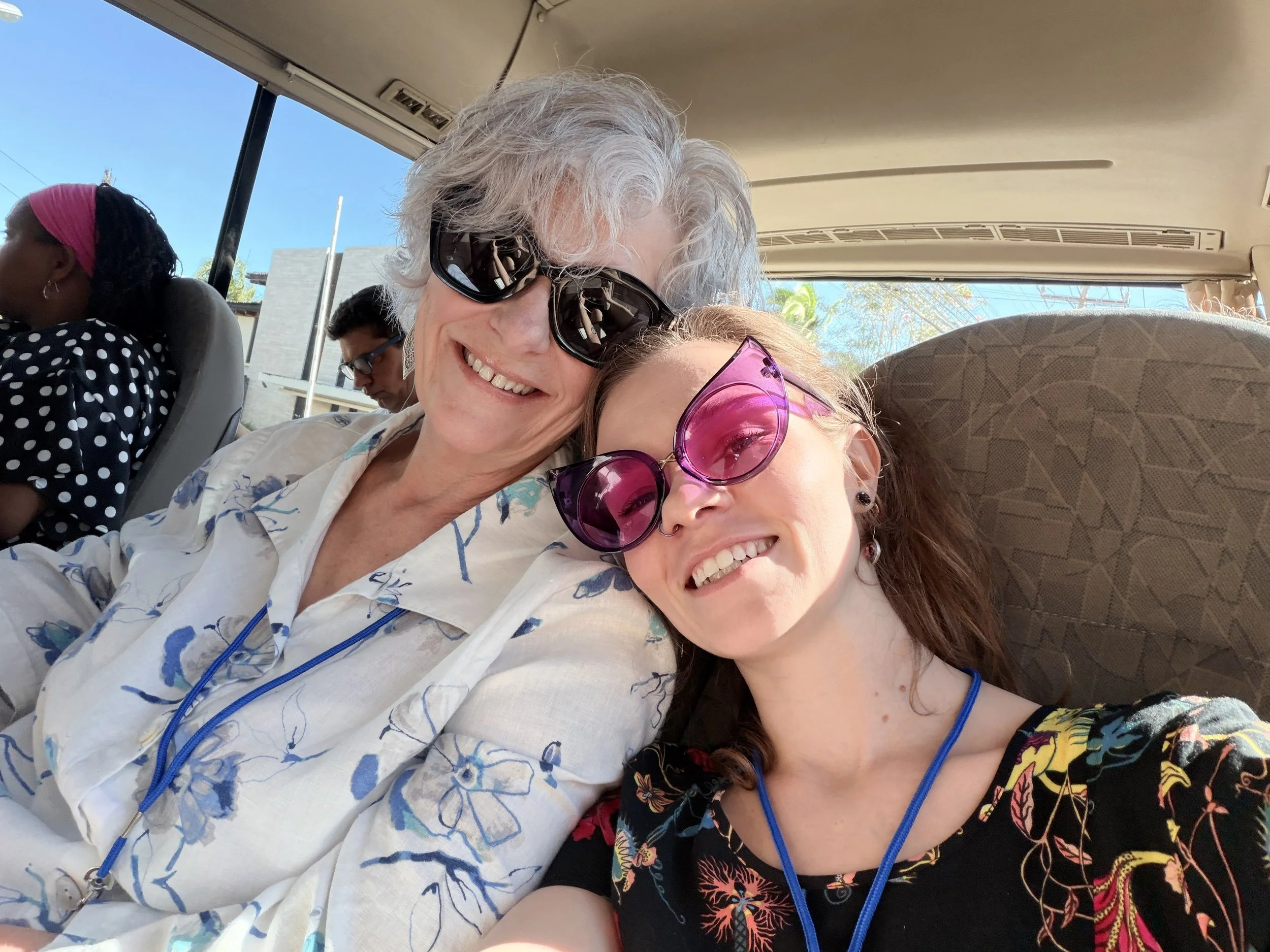 Two women and two men sitting inside a bus or van, smiling and wearing sunglasses. The women are in the foreground, one with gray curly hair and the other with long reddish-brown hair, both smiling brightly. The background shows a woman with a pink headscarf and sunglasses, and a man with dark hair and glasses, all enjoying a sunny day.