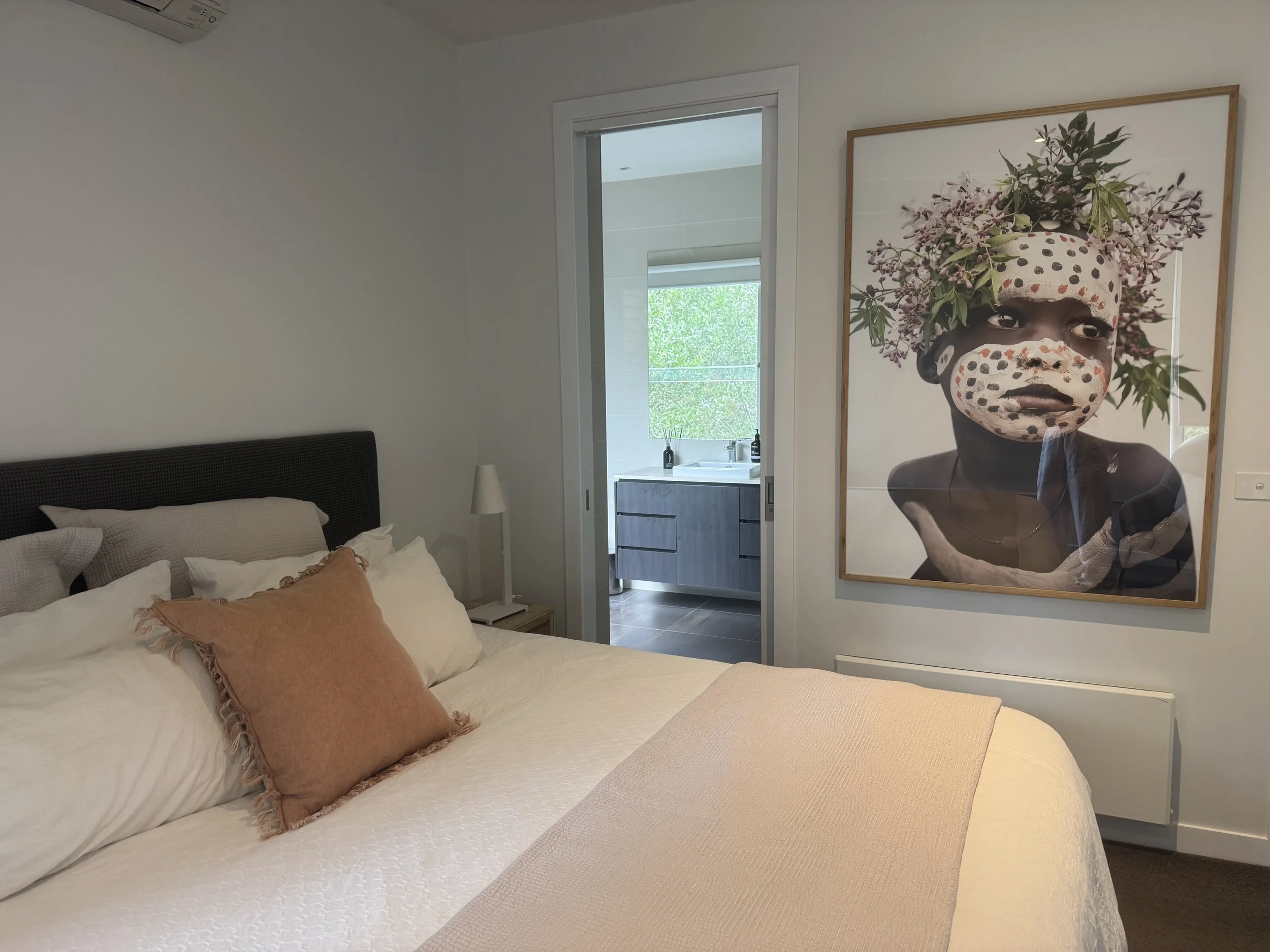 A bedroom with a bed, pillows, and a bedside lamp. There is a large framed artwork of a child with painted face and a floral headdress hanging on the wall. An open door reveals a bathroom with a sink and window outside.